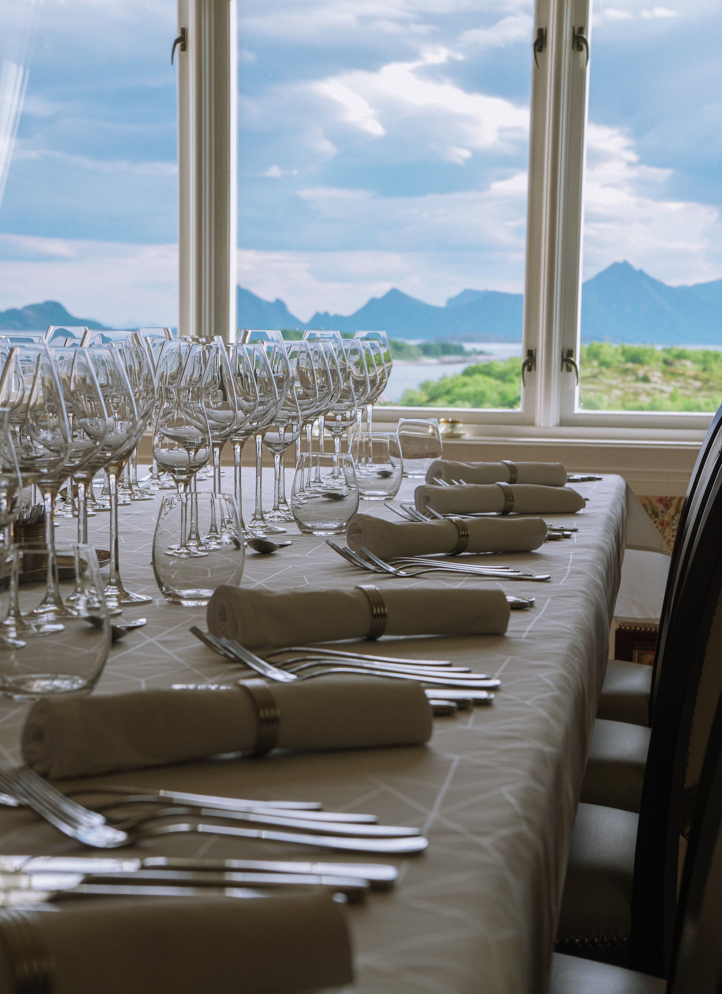 Empty table set for a meal with wine glasses, water glasses, rolled napkins, forks, knives, and spoons, positioned next to a large window showing a mountain landscape with a lake and trees, with a partly cloudy sky.
