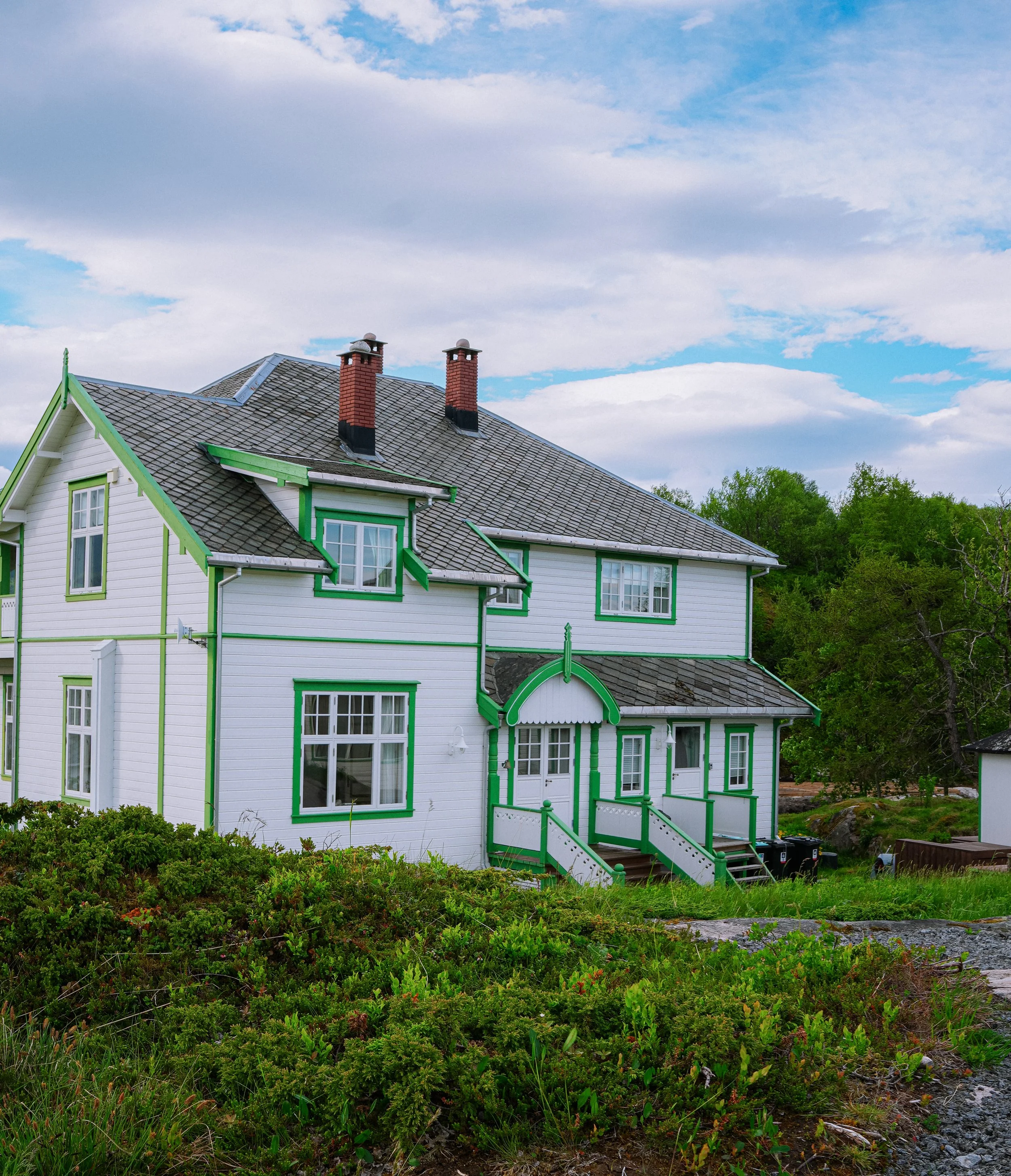 A white house with green trim and multiple chimneys on the roof, surrounded by green vegetation and trees, under a partly cloudy sky.