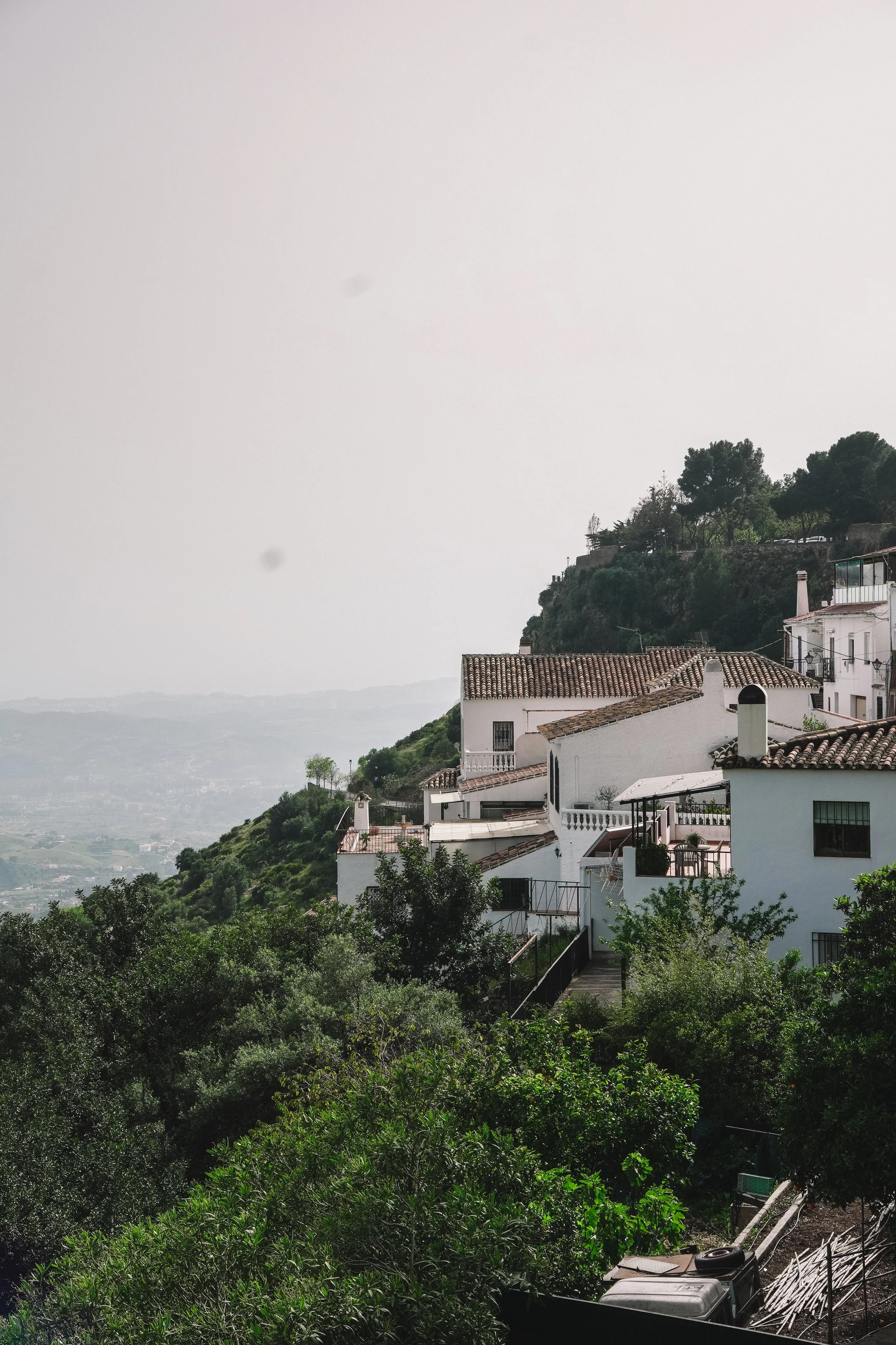 A hillside view featuring white Mediterranean-style houses with terracotta tiled roofs, surrounded by lush green trees and vegetation, overlooking a distant valley under an overcast sky.
