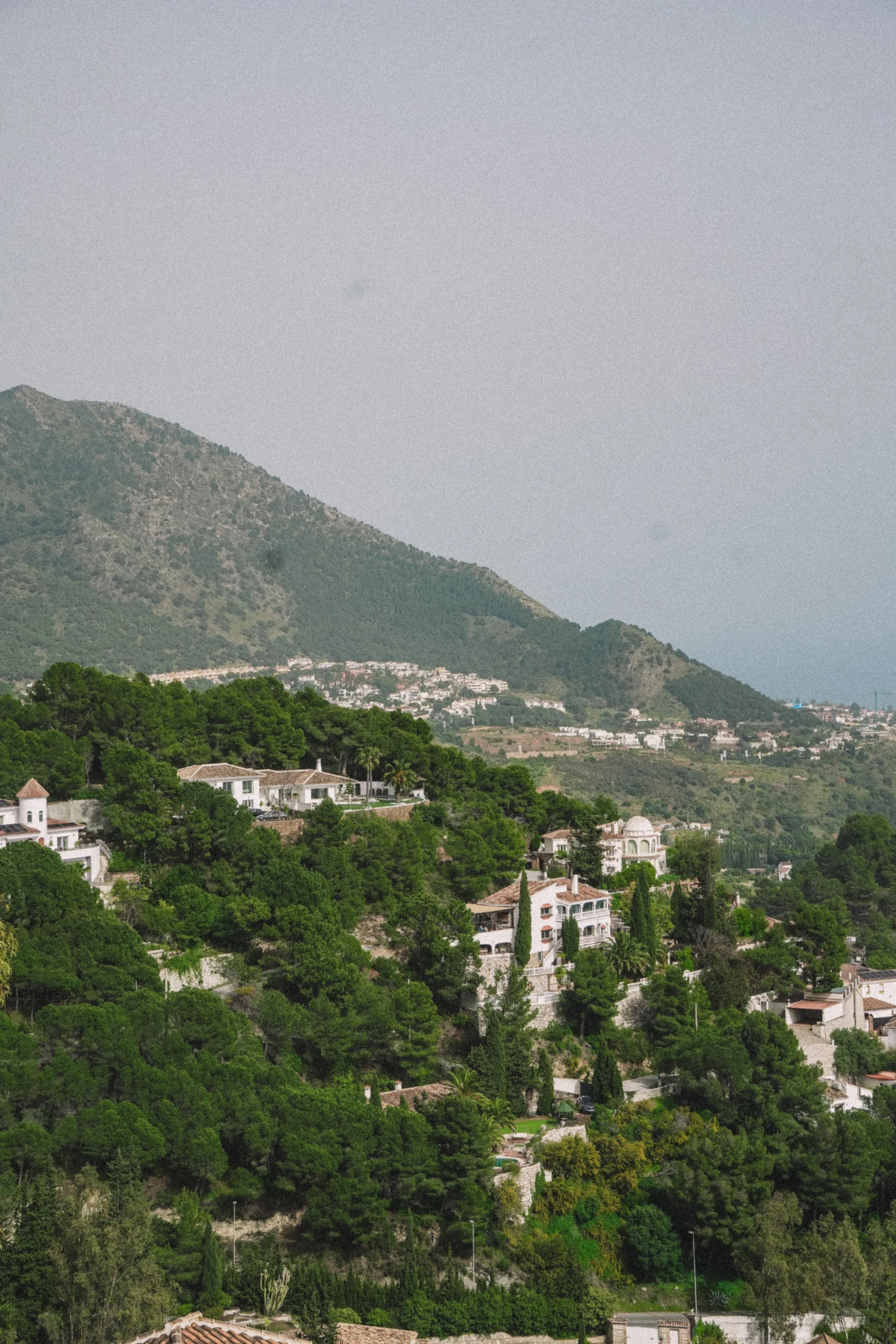 Hilly landscape with scattered homes and lush greenery, mountains in the background under a partly cloudy sky.