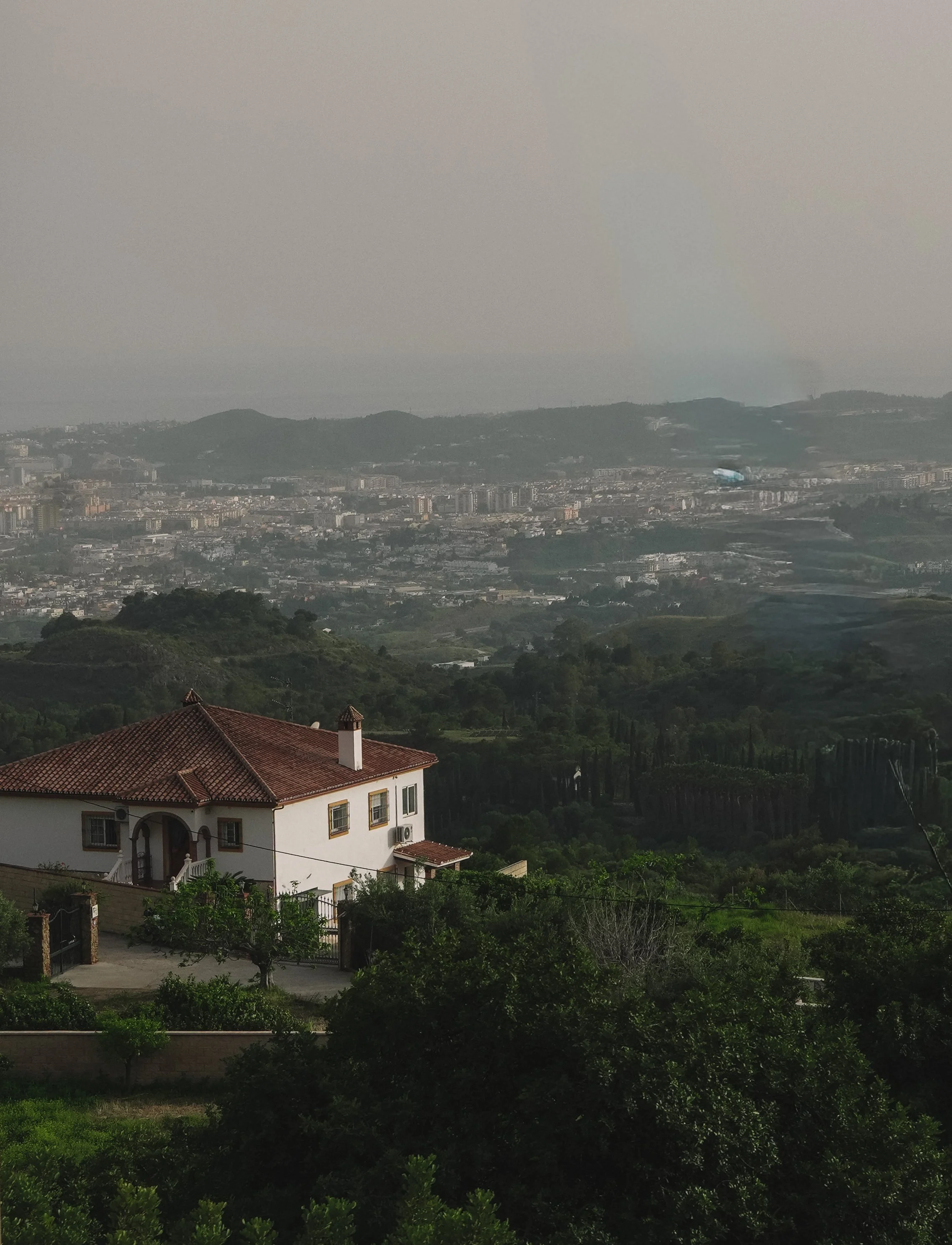 A scenic view of a hilly landscape with a city in the background, featuring a house with a red-tiled roof in the foreground, and mountains and a hazy sky in the distance.