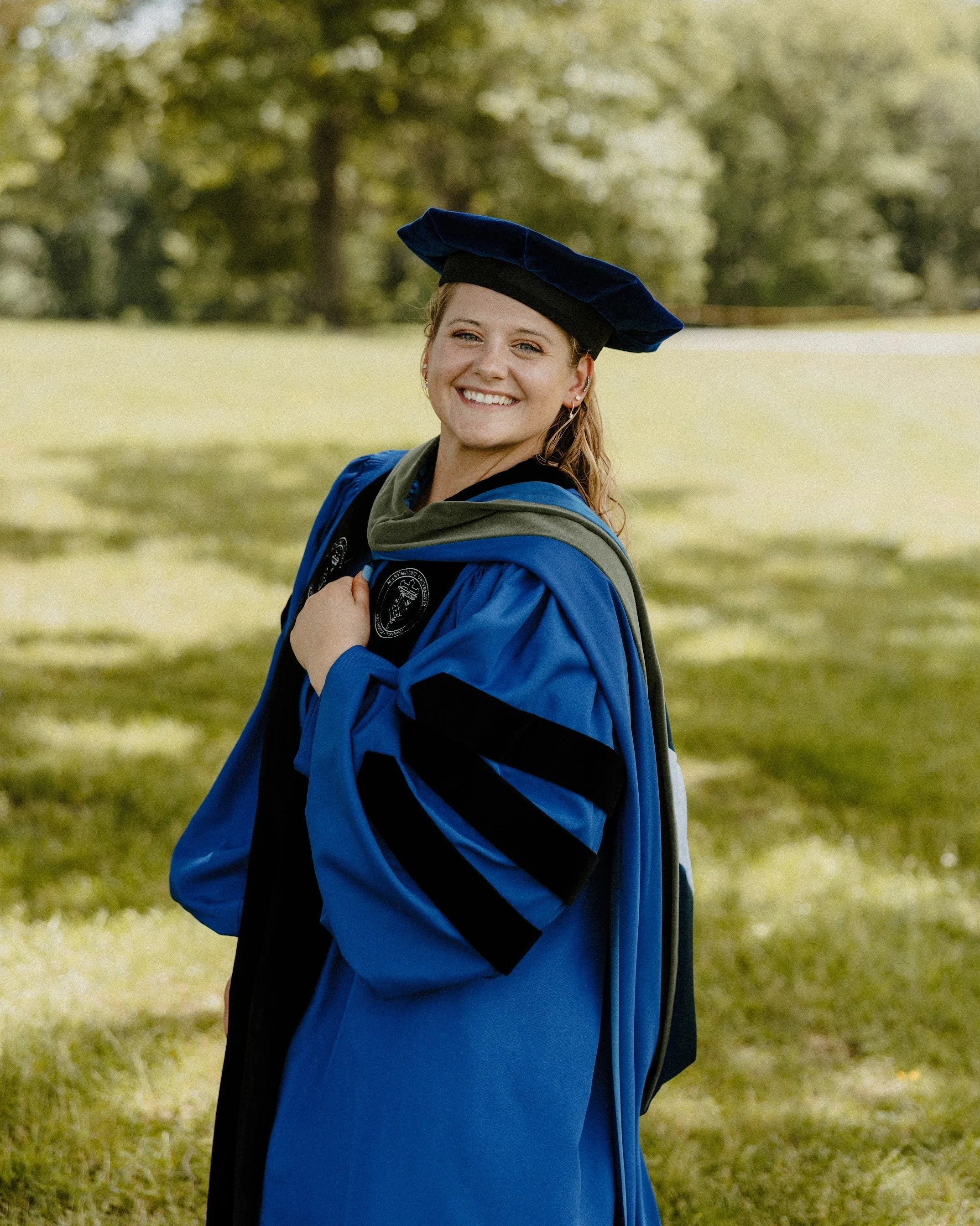 A young woman in a blue graduation gown and cap stands outdoors on grass with trees in the background, smiling and looking at the camera.