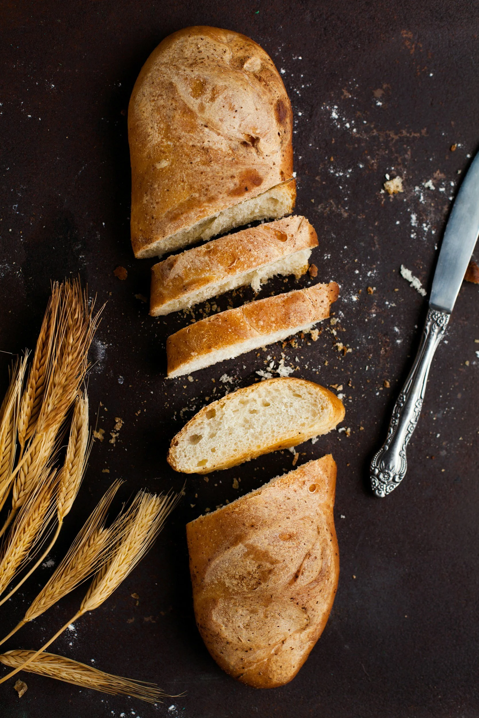 Broken loaf of bread on a dark surface, with wheat stalks nearby, and a knife next to the bread.