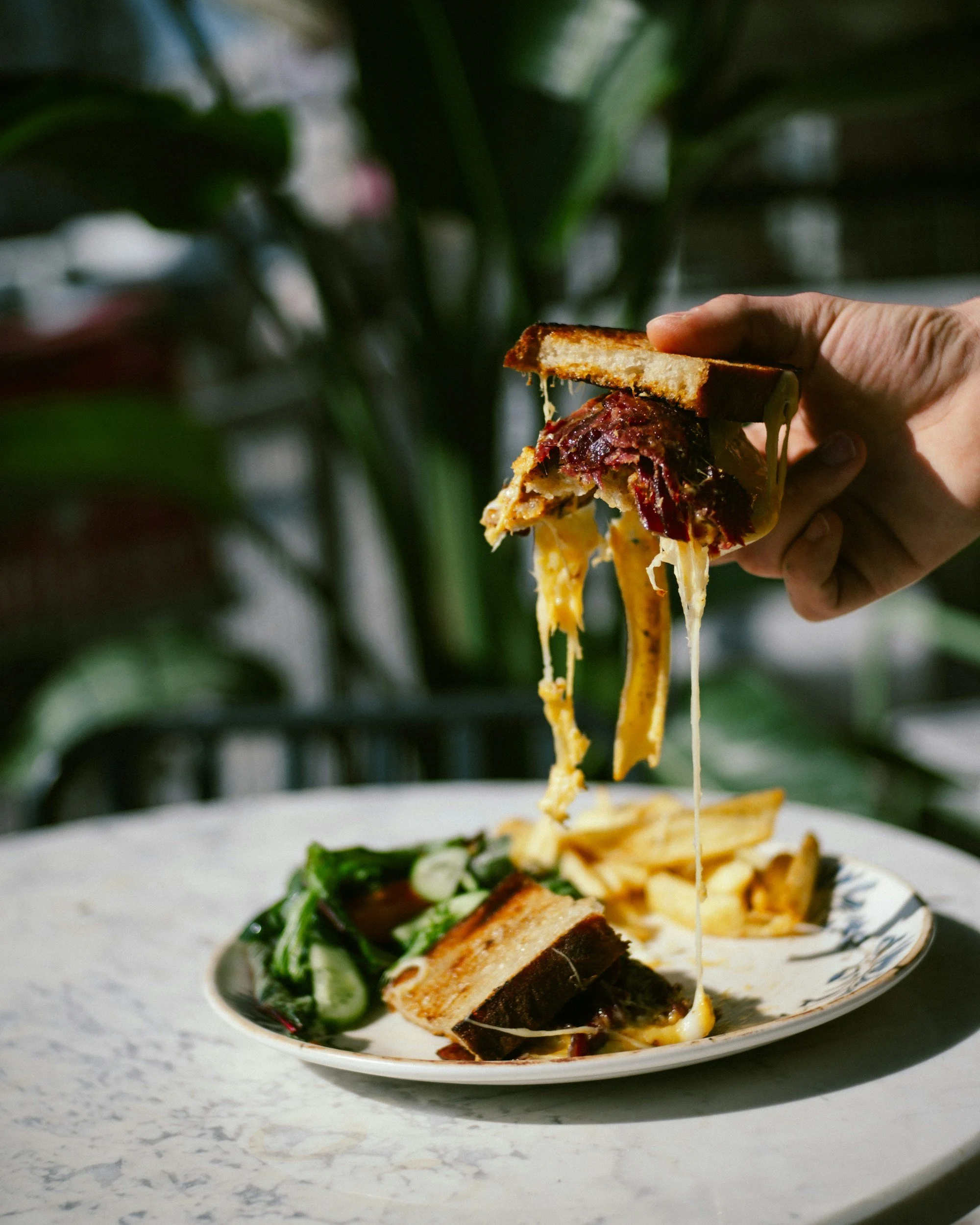 A plate of food with pasta, grilled chicken, spinach, and a slice of bread. Someone is holding a slice of bread with melted cheese and toppings, lifting it above the plate.