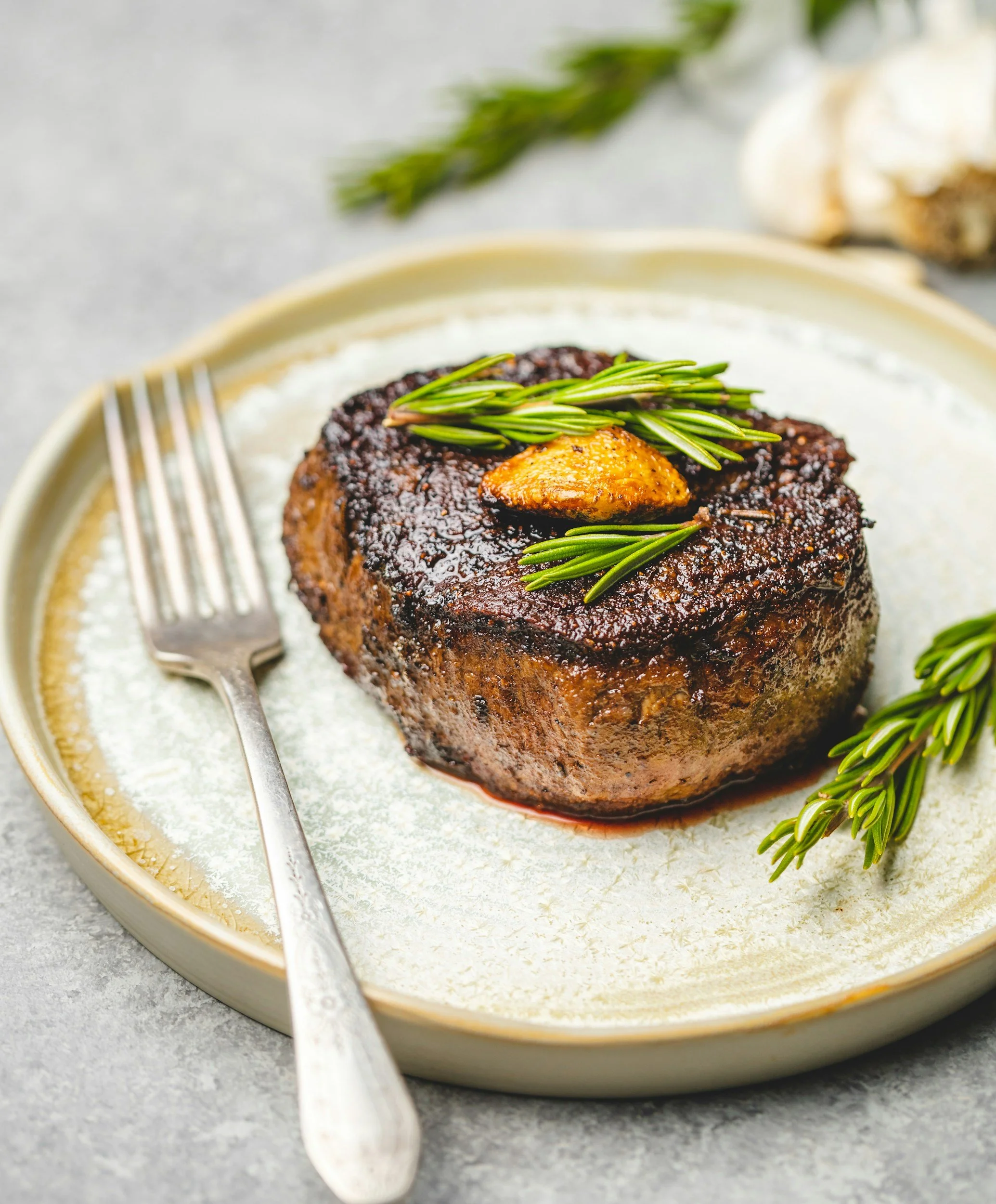 Close-up of a grilled beef patty topped with a roasted garlic clove and sprigs of fresh rosemary on a white plate with a fork and on a gray surface.