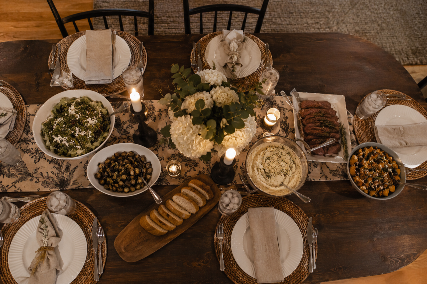 A family dinner table with a floral centerpiece, various dishes including salad, roasted vegetables, sliced bread, and meats, set for six people with candles and tableware.