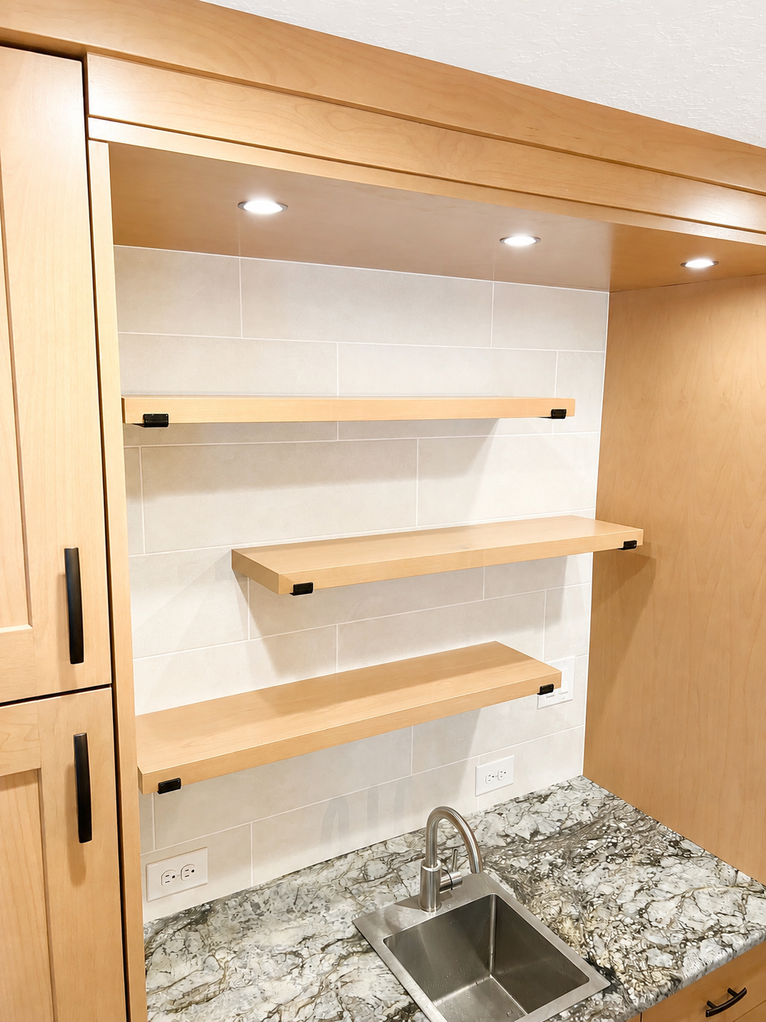 Kitchen with wooden cabinets, granite countertop, sink, beige tile backsplash, and three wooden shelves with black brackets under three ceiling lights.