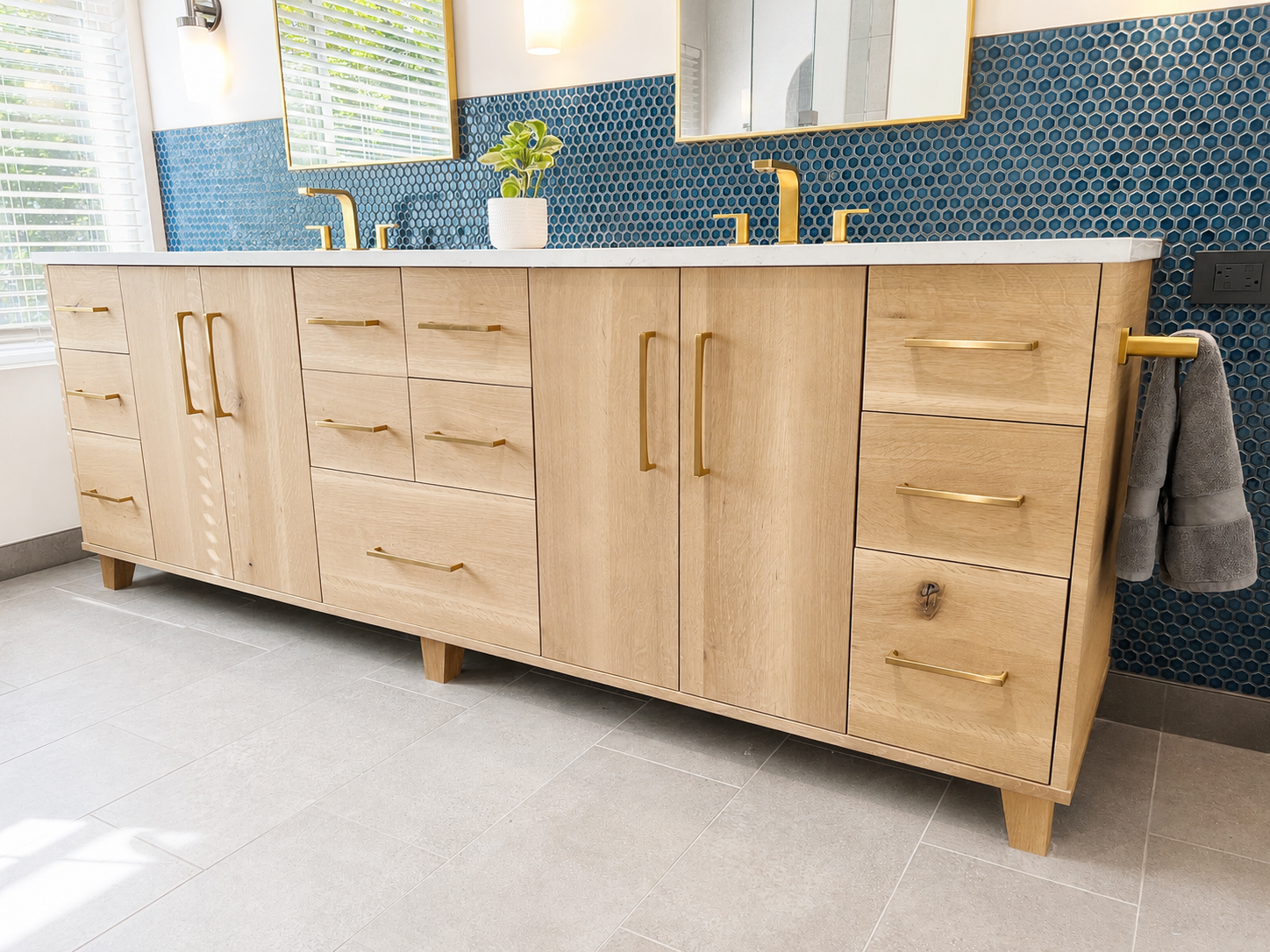 Wooden bathroom vanity with gold handles, a white countertop, two gold faucets, a mirror, and a blue mosaic tile backsplash with a potted plant.