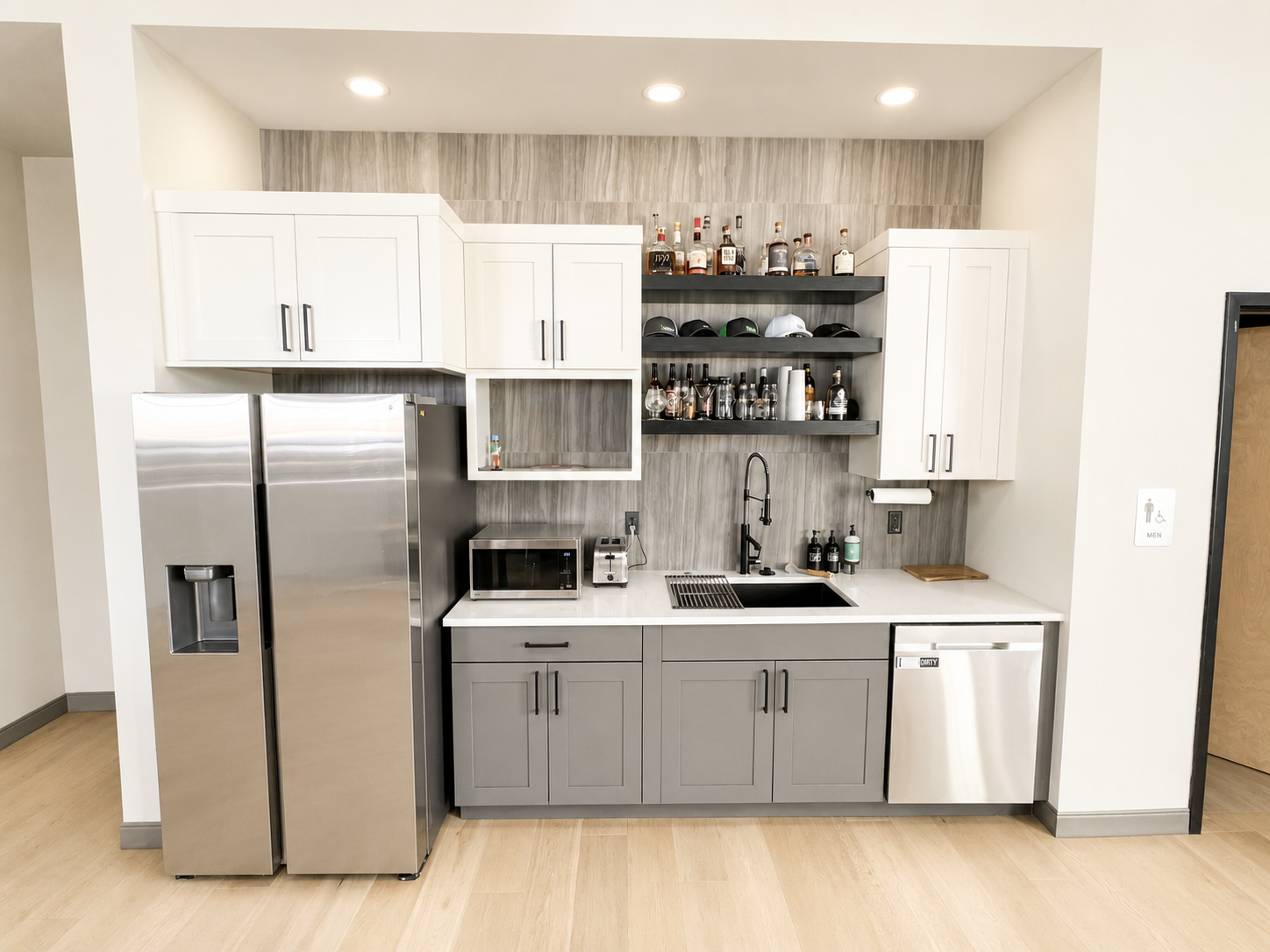 Modern kitchen with gray lower cabinets, white upper cabinets, stainless steel refrigerator, microwave, and small dishwasher, open shelving with liquor bottles, cups, and caps, black faucet, and wooden floor.