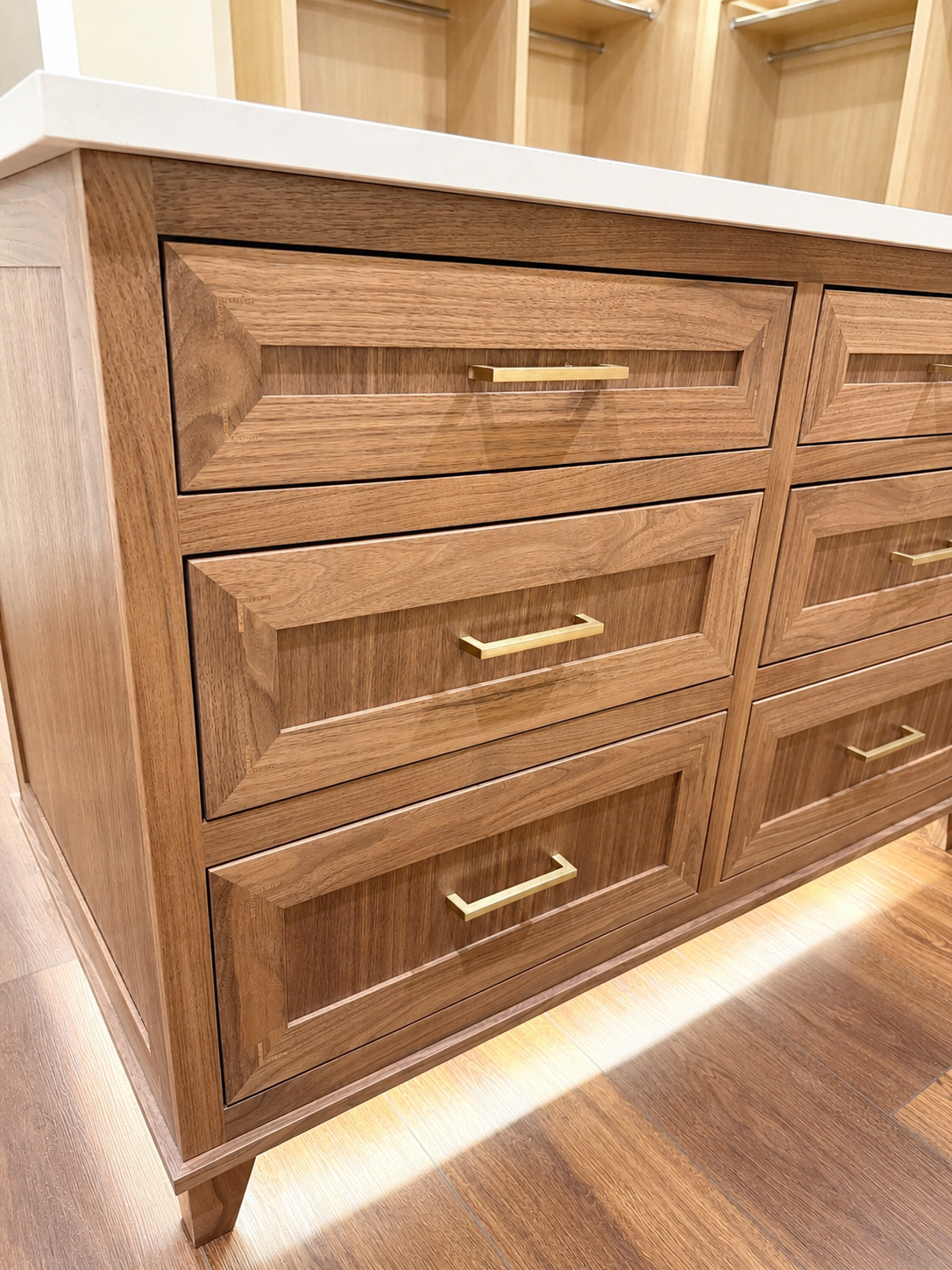 A wooden dresser with six drawers and gold handles, situated in a room with wooden flooring and a partially visible wooden shelving unit in the background.