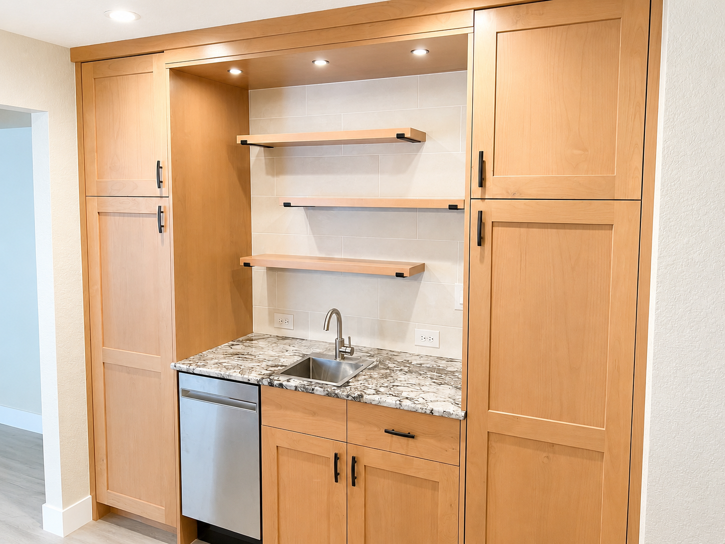 Small kitchenette with wooden cabinets, granite countertop, a small sink, and an under-counter dishwasher. Two open shelves are mounted on the tiled wall above the sink.