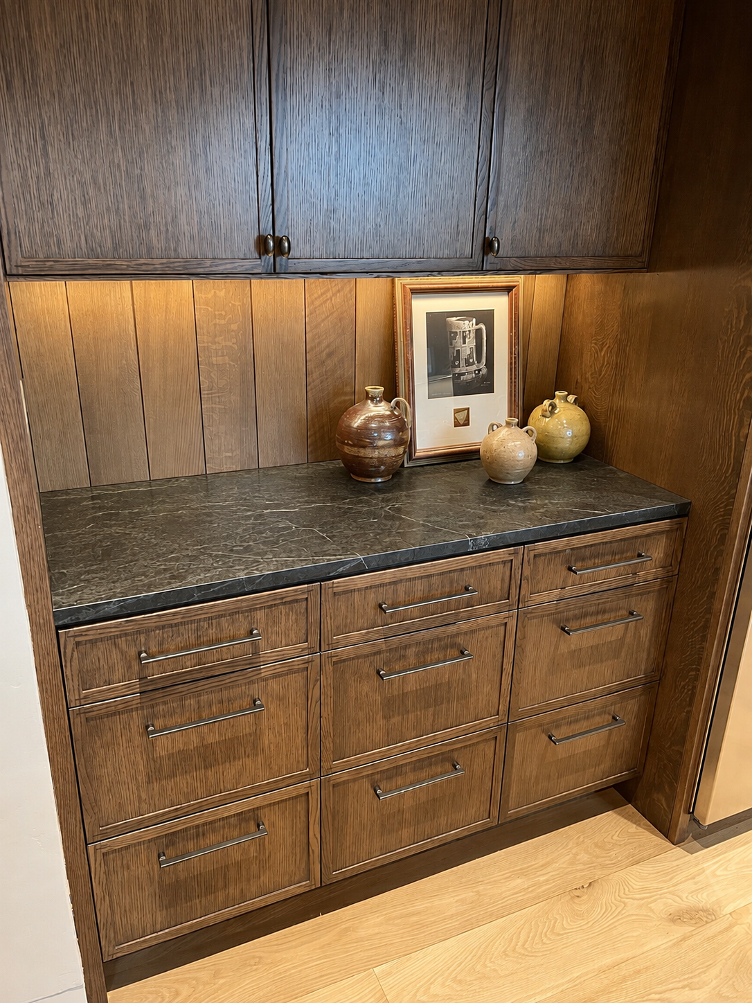 A wooden kitchen cabinet with a black marble countertop, three ceramic vases, and framed artwork on top.