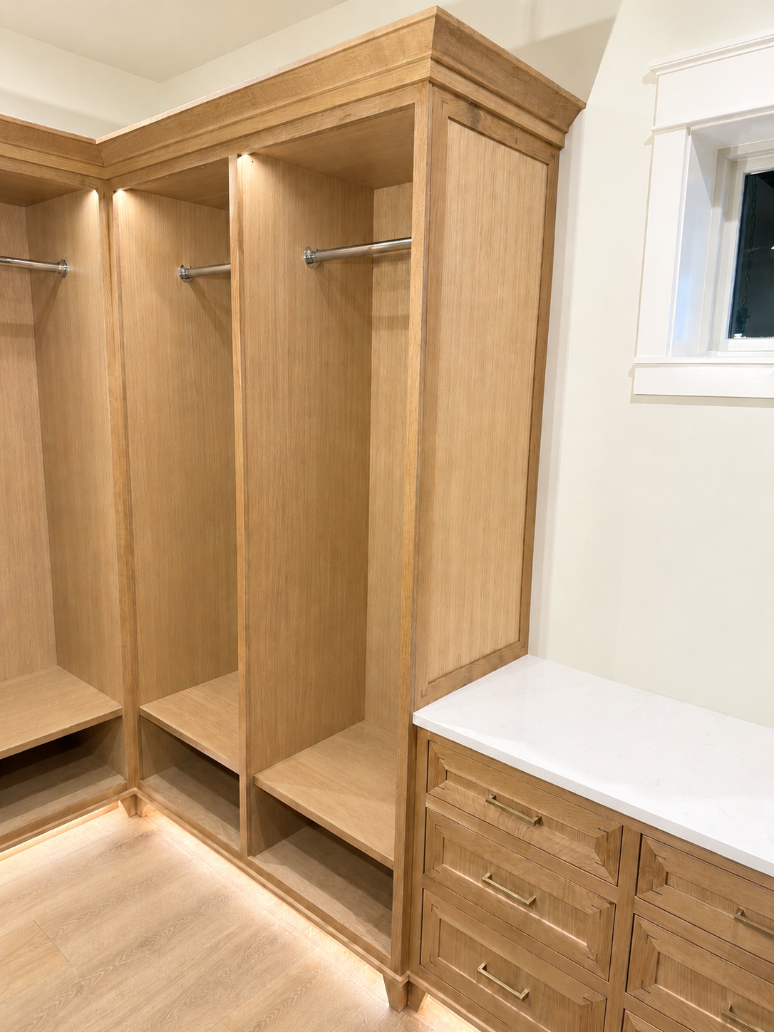 Empty wooden walk-in closet with built-in drawers and hanging rods, featuring recessed lighting and a window.