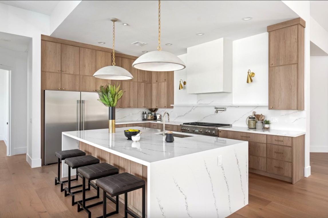 Modern kitchen with white marble countertops, wooden cabinets, stainless steel refrigerator, and four barstools at the island, with pendant lights, wall-mounted sconces, and decor items.