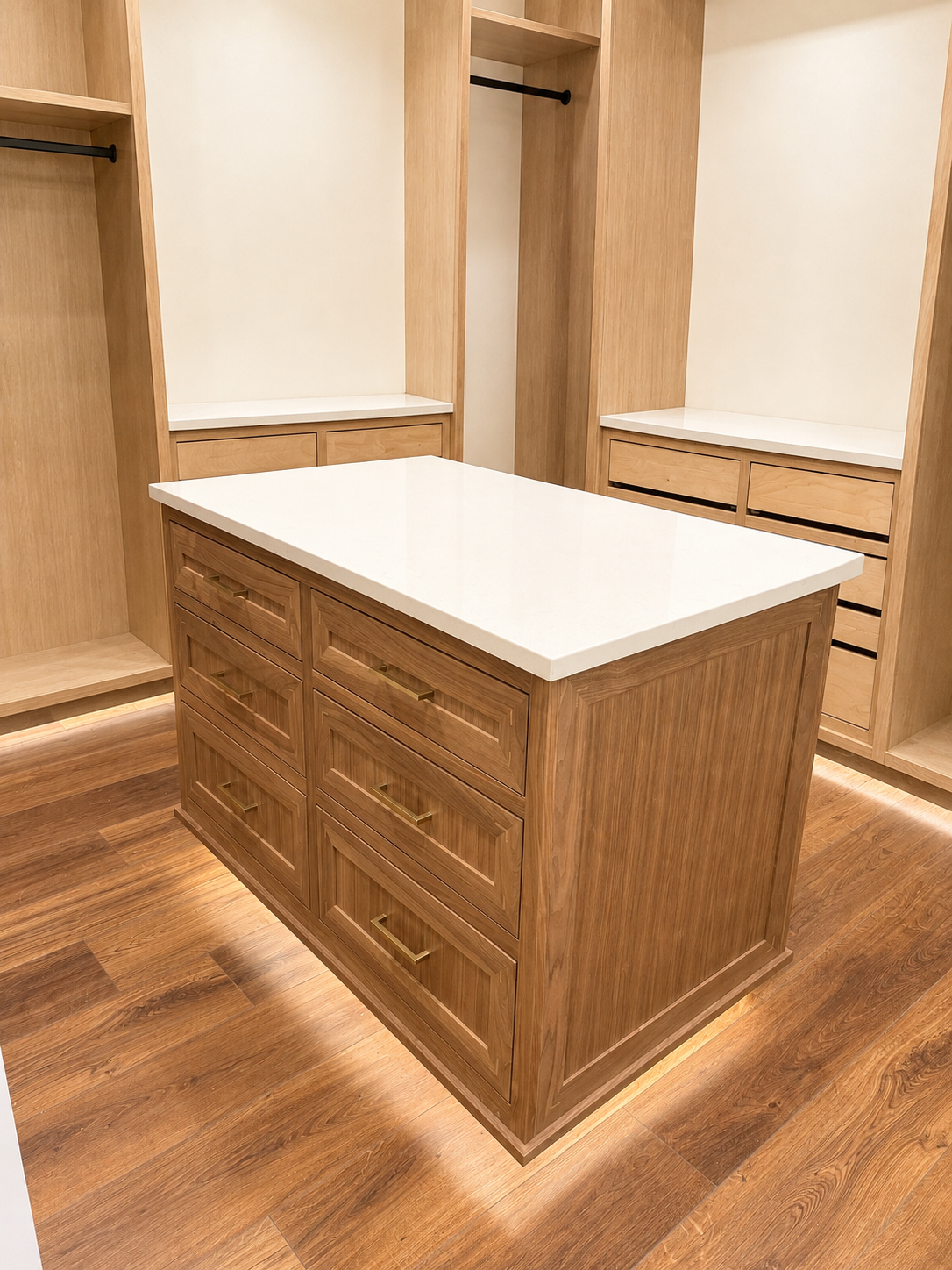 A wooden walk-in closet with a central island featuring six drawers with gold handles, open shelving units, and hanging rods, illuminated by under-cabinet lighting.