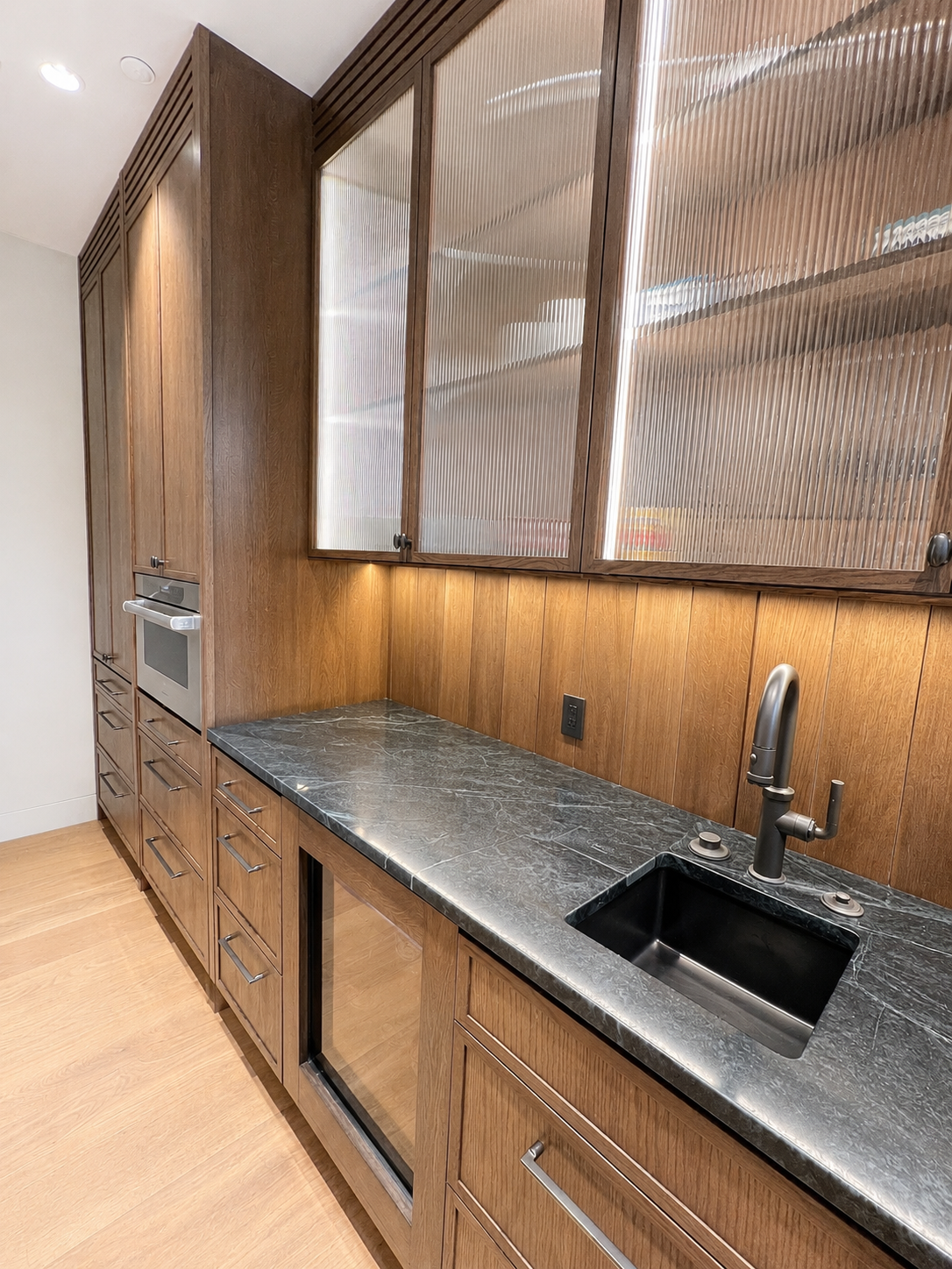 Modern kitchen with wooden cabinets, a black marble countertop, a built-in oven, a small refrigerator, and a black sink with a silver faucet.