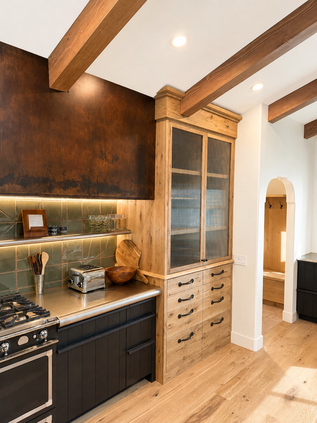 Kitchen with wooden beams ceiling, black lower cabinets, a stovetop, a stainless steel countertop, a wooden bowl, glassware, a toaster, and a wooden cabinet with glass doors.
