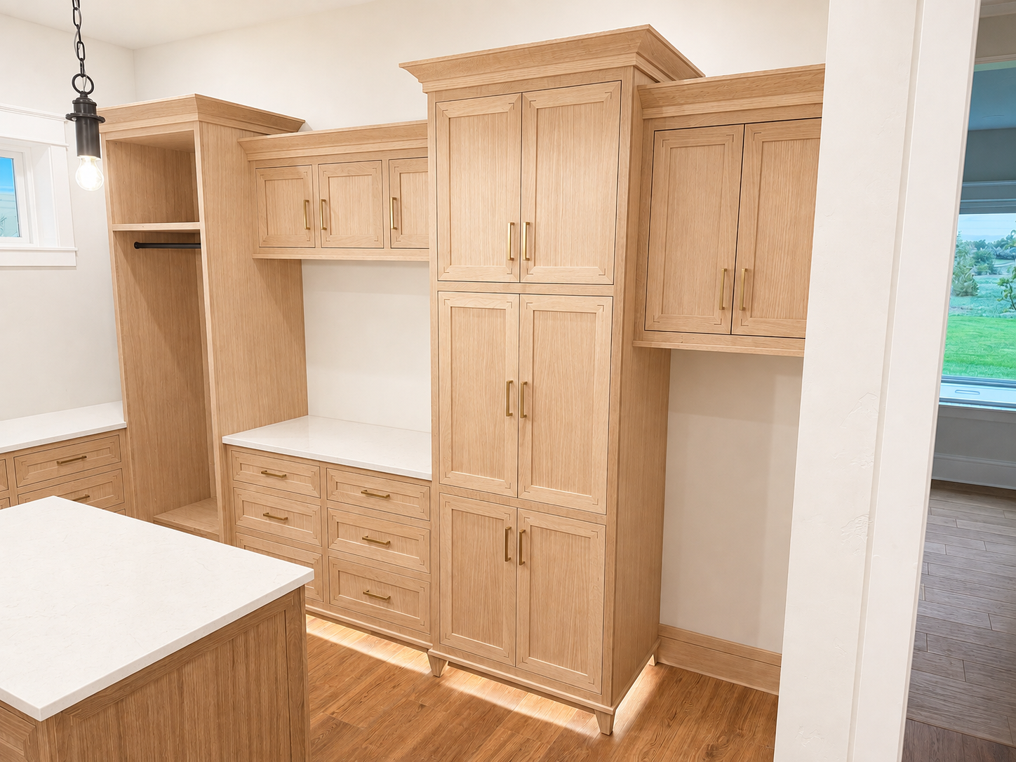 Built-in wooden kitchen cabinetry with multiple cabinets and drawers, a small countertop space, and an open section with a hanging rod, with a light fixture hanging from the ceiling and a window showing a green outdoor landscape.