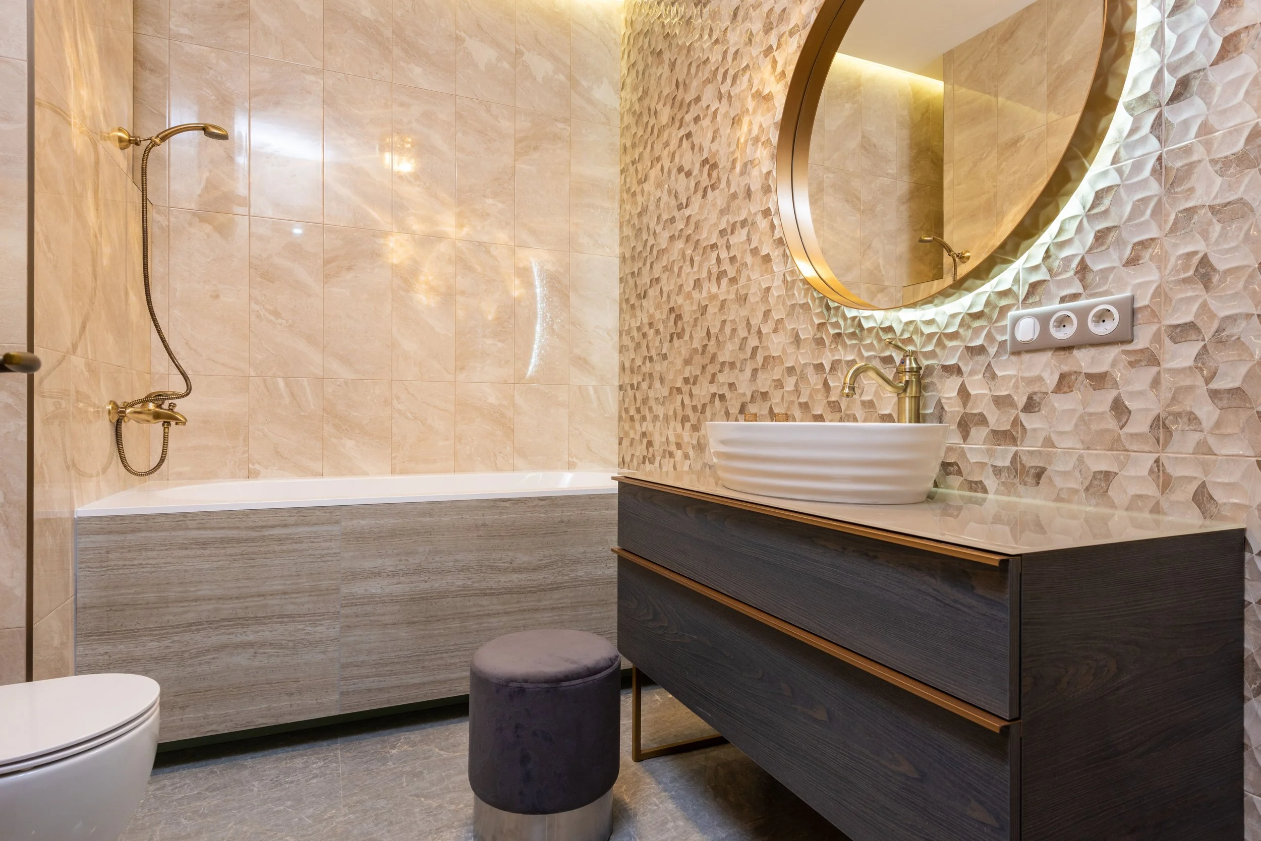 Modern bathroom with beige tiled walls and floor, a round mirror with backlight, a vessel sink with gold faucet, a built-in bathtub with beige tiles, a small round gray stool, and a dual electrical outlet.
