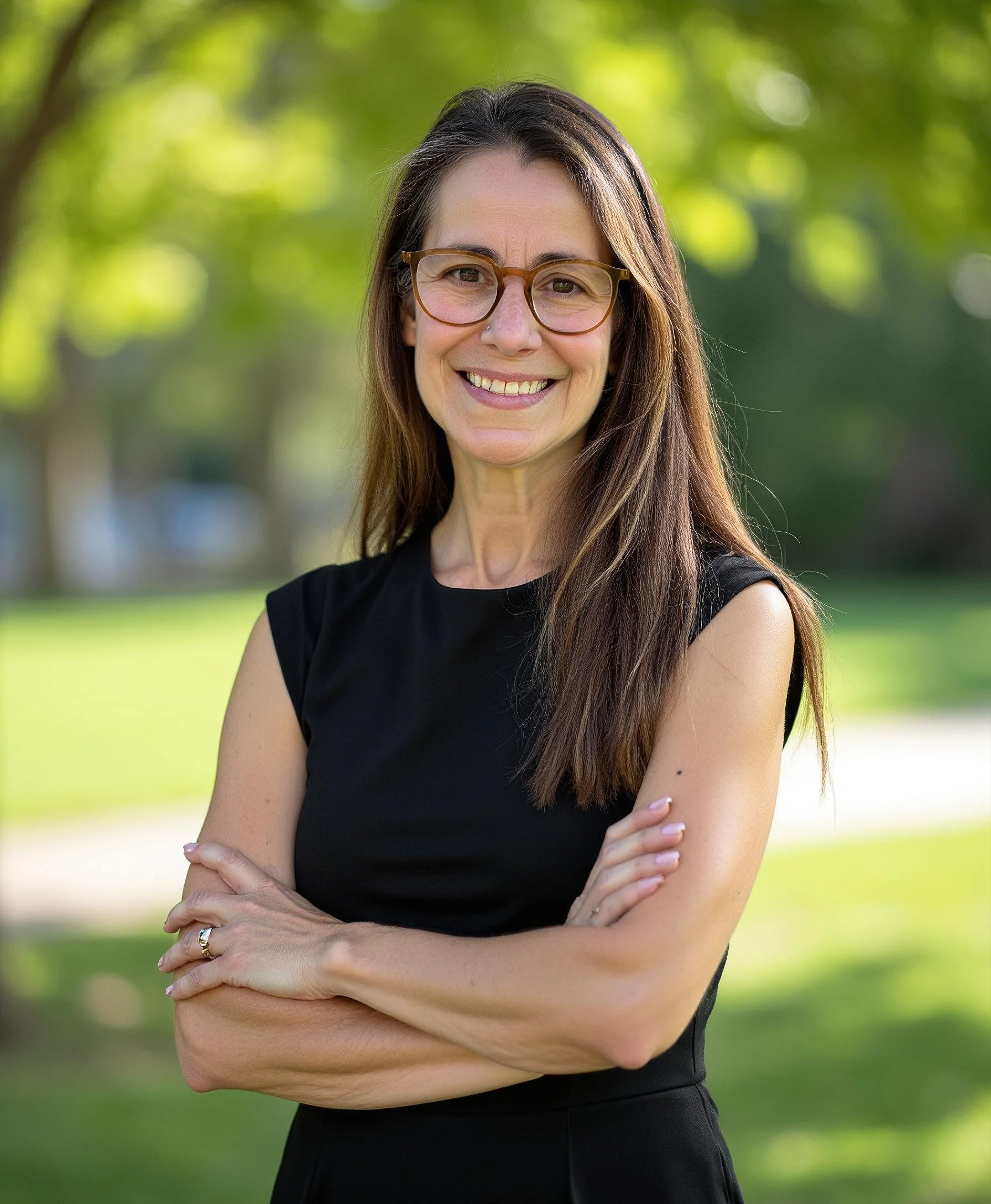 A woman with long brown hair, glasses, and a black sleeveless top standing outdoors with arms crossed, smiling at the camera, with green trees and sunlight in the background.