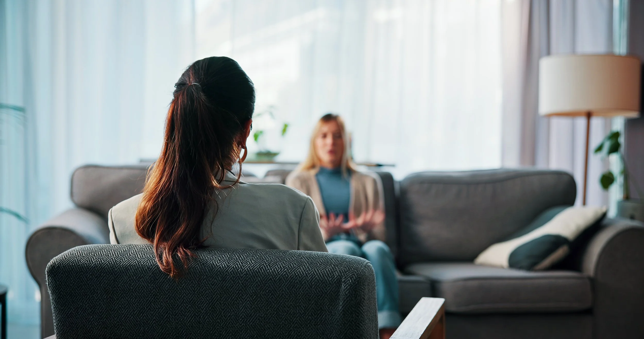 Two women are sitting and talking in a cozy living room with large windows and natural light, surrounded by plants and cozy furniture.