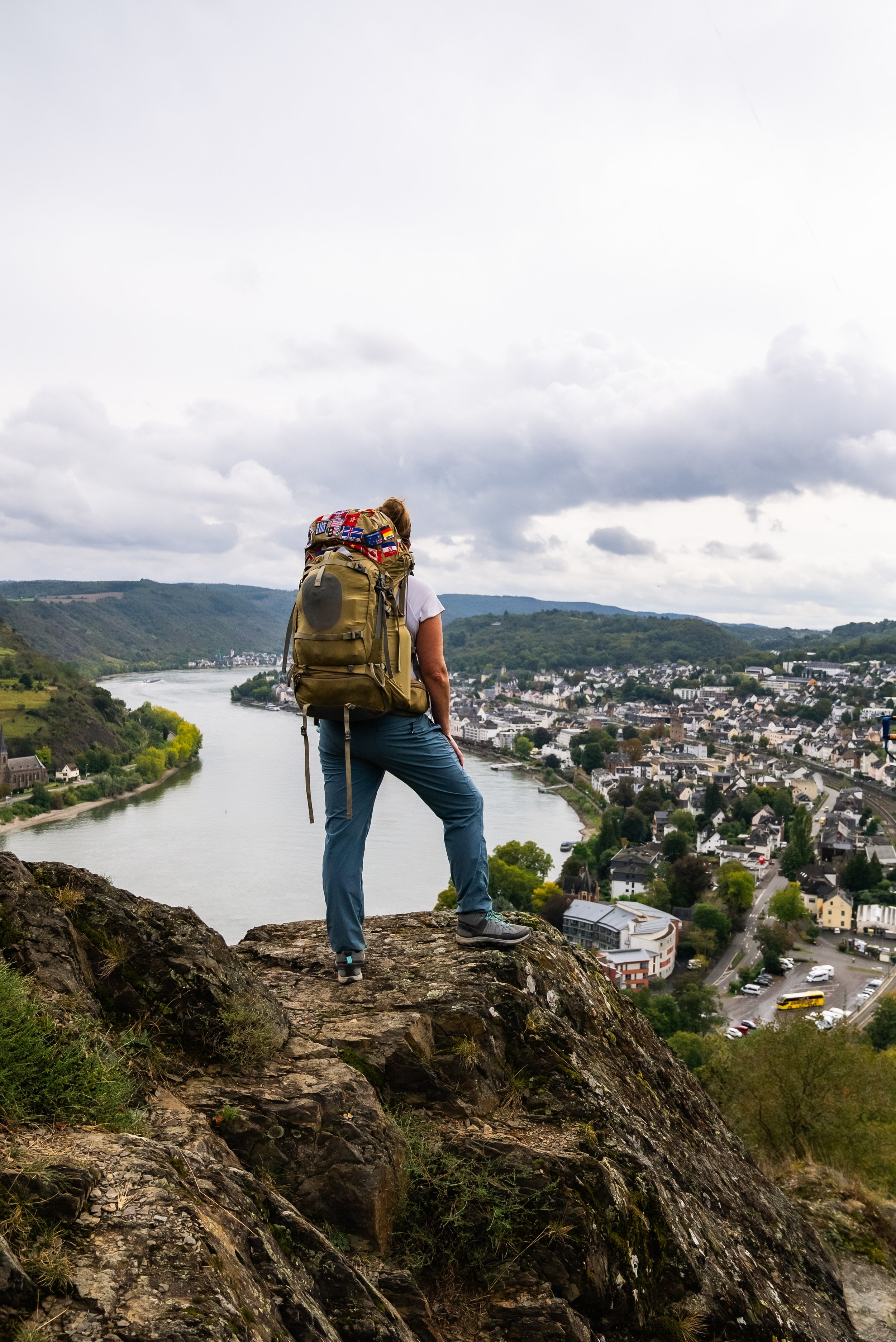 Person with long hair and hiking backpack standing on rocky ledge overlooking a town and river, cloudy sky