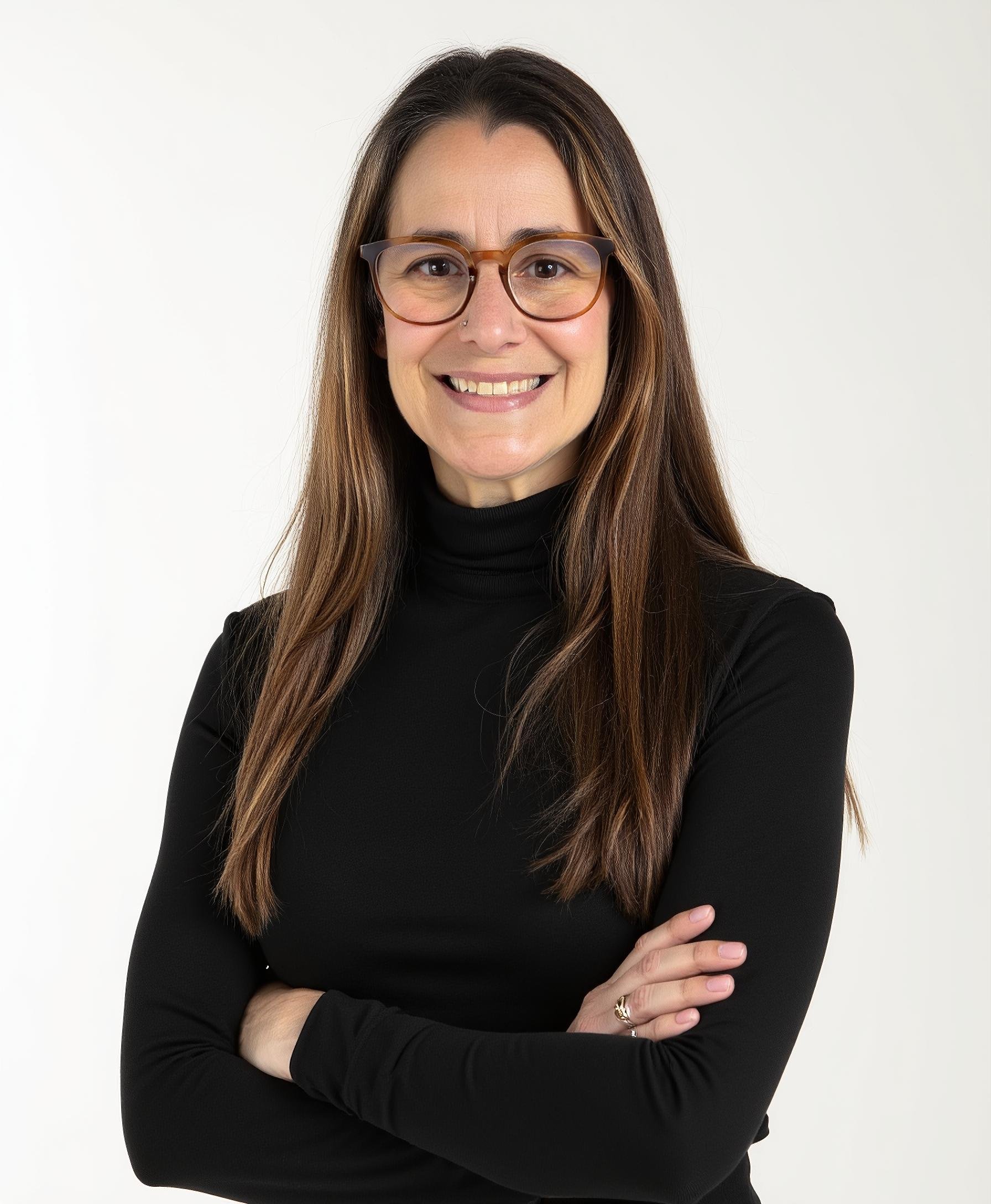 A woman with long brown hair wearing glasses and a black turtleneck, smiling with her arms crossed against a plain white background.