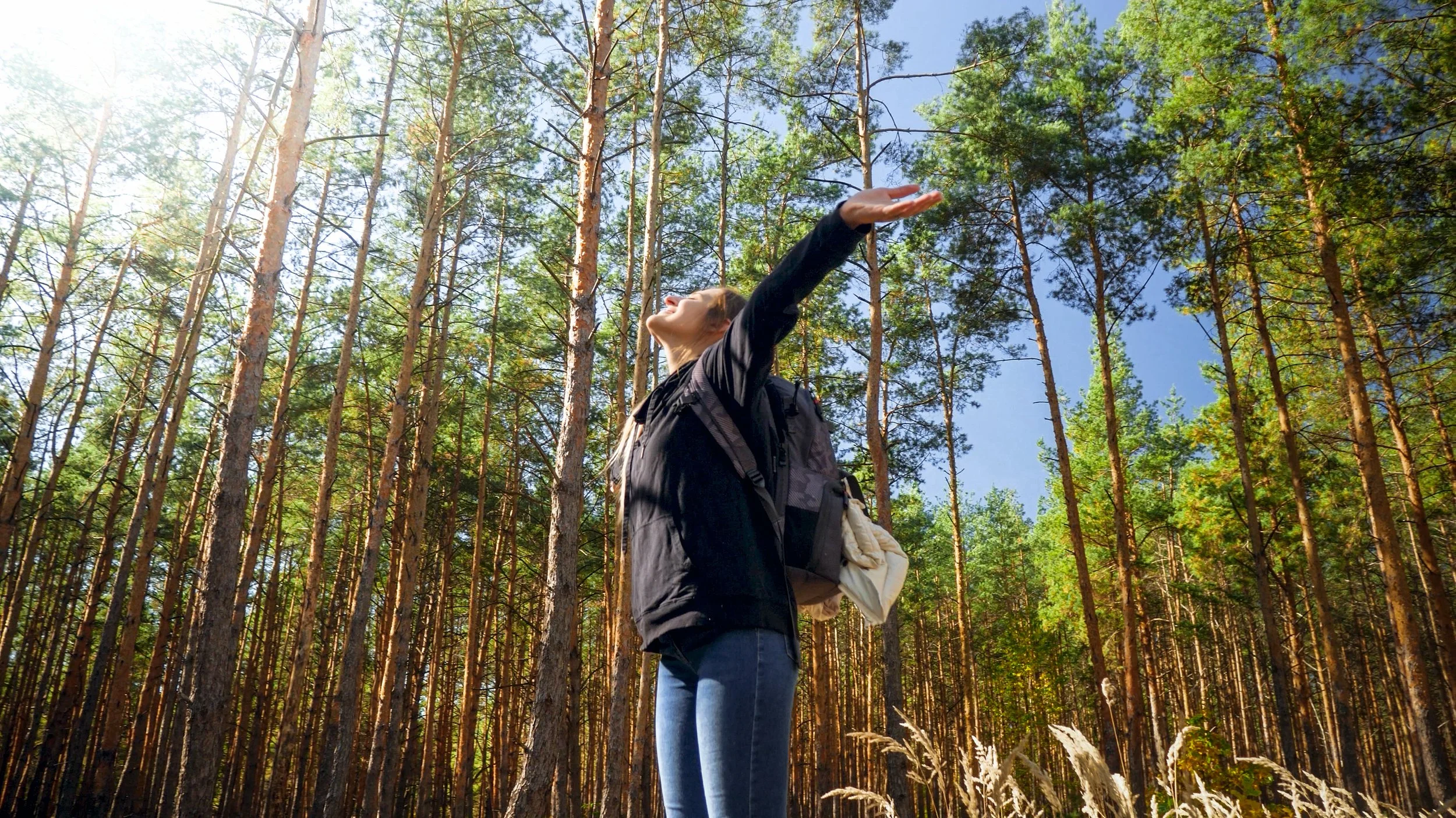 A woman in a black jacket standing among tall pine trees with her arms raised toward the sky, smiling and enjoying the outdoors on a sunny day.