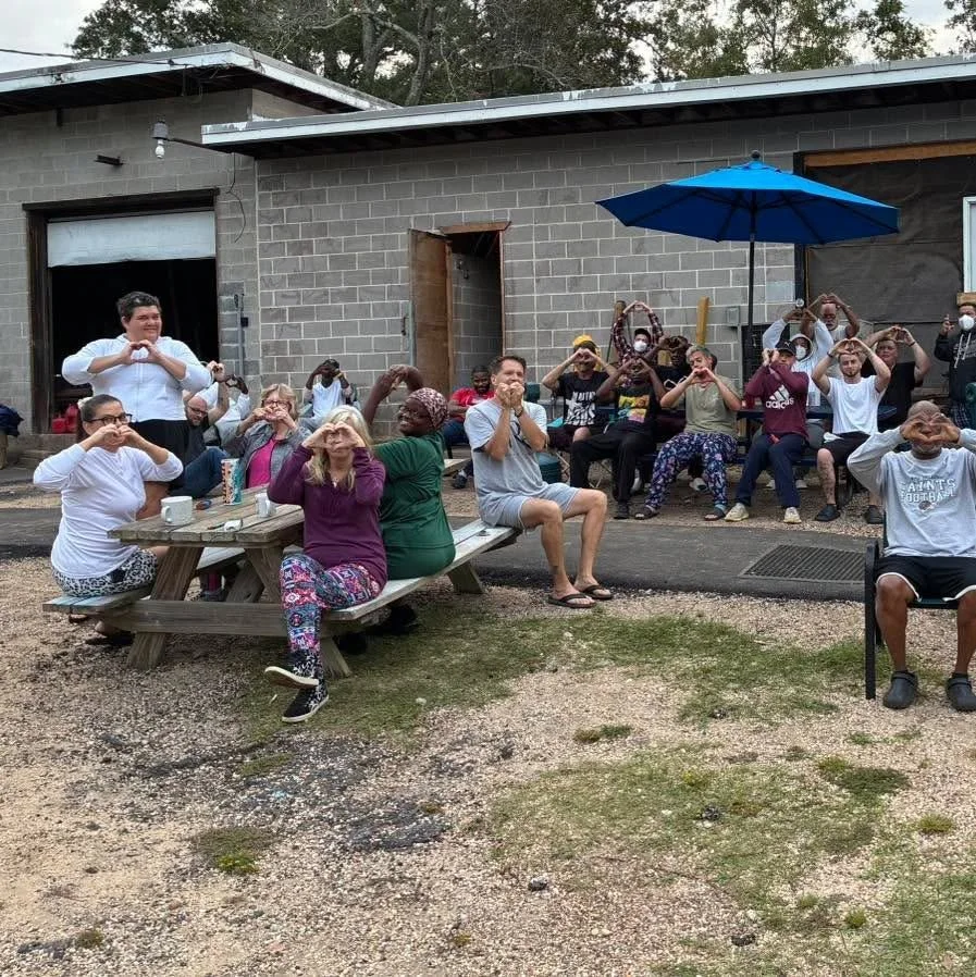 Group of adults and children sitting outside, many making heart shapes with their hands or forming hearts with their fingers, in front of a residential building with a blue umbrella and wooden picnic tables.