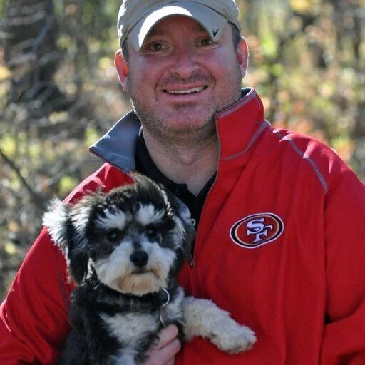 A man smiling outdoors holding a small black and white puppy, both facing the camera.
