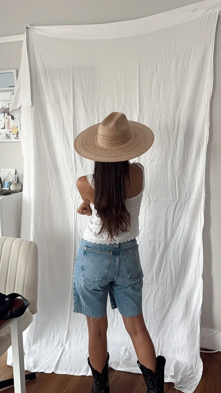A woman standing with her back to the camera, wearing a large straw hat, white sleeveless top, denim shorts, and black cowboy boots, in front of a crumpled white backdrop.