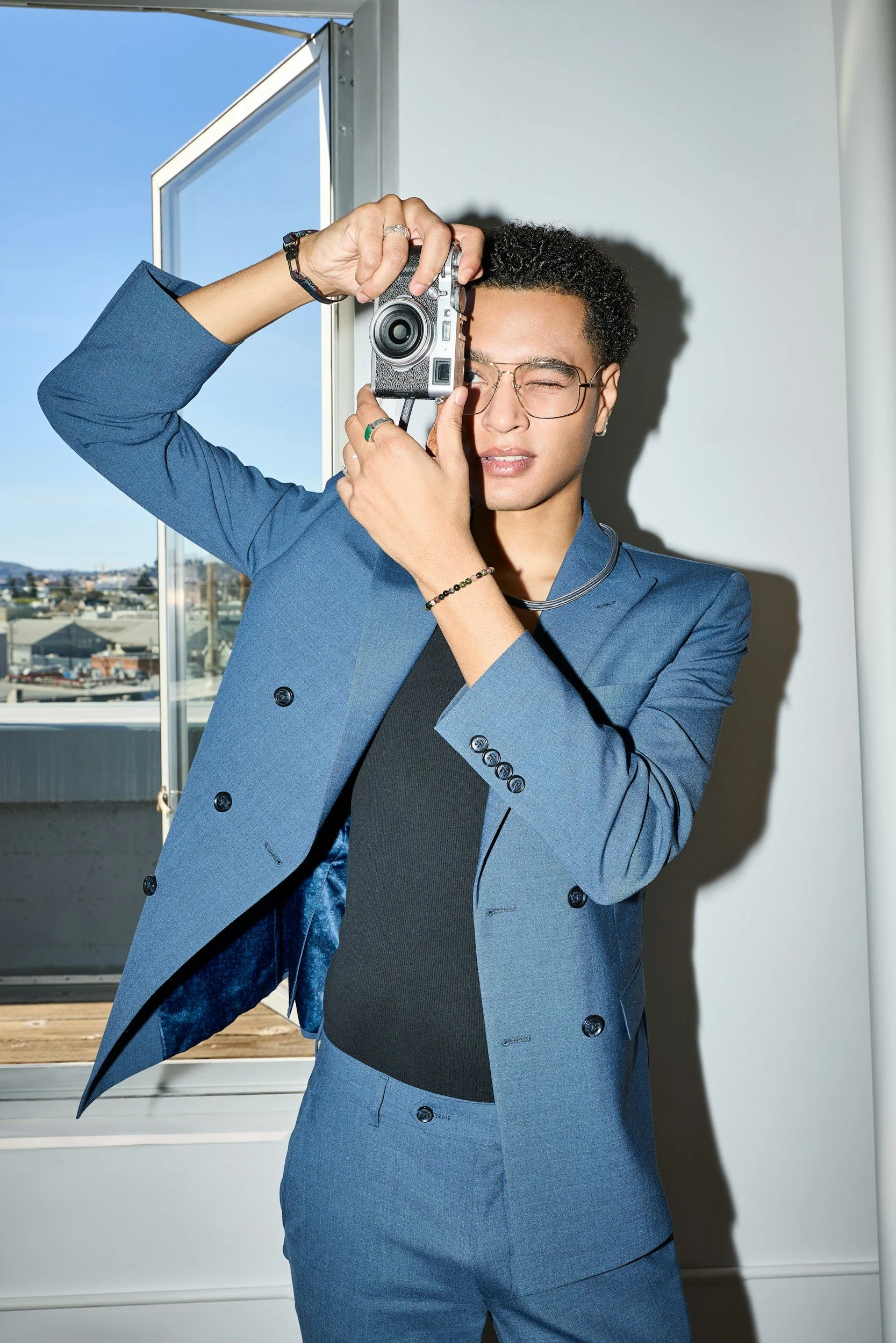 Person in a blue suit taking a photo or looking through a camera indoors near a window with a cityscape view.
