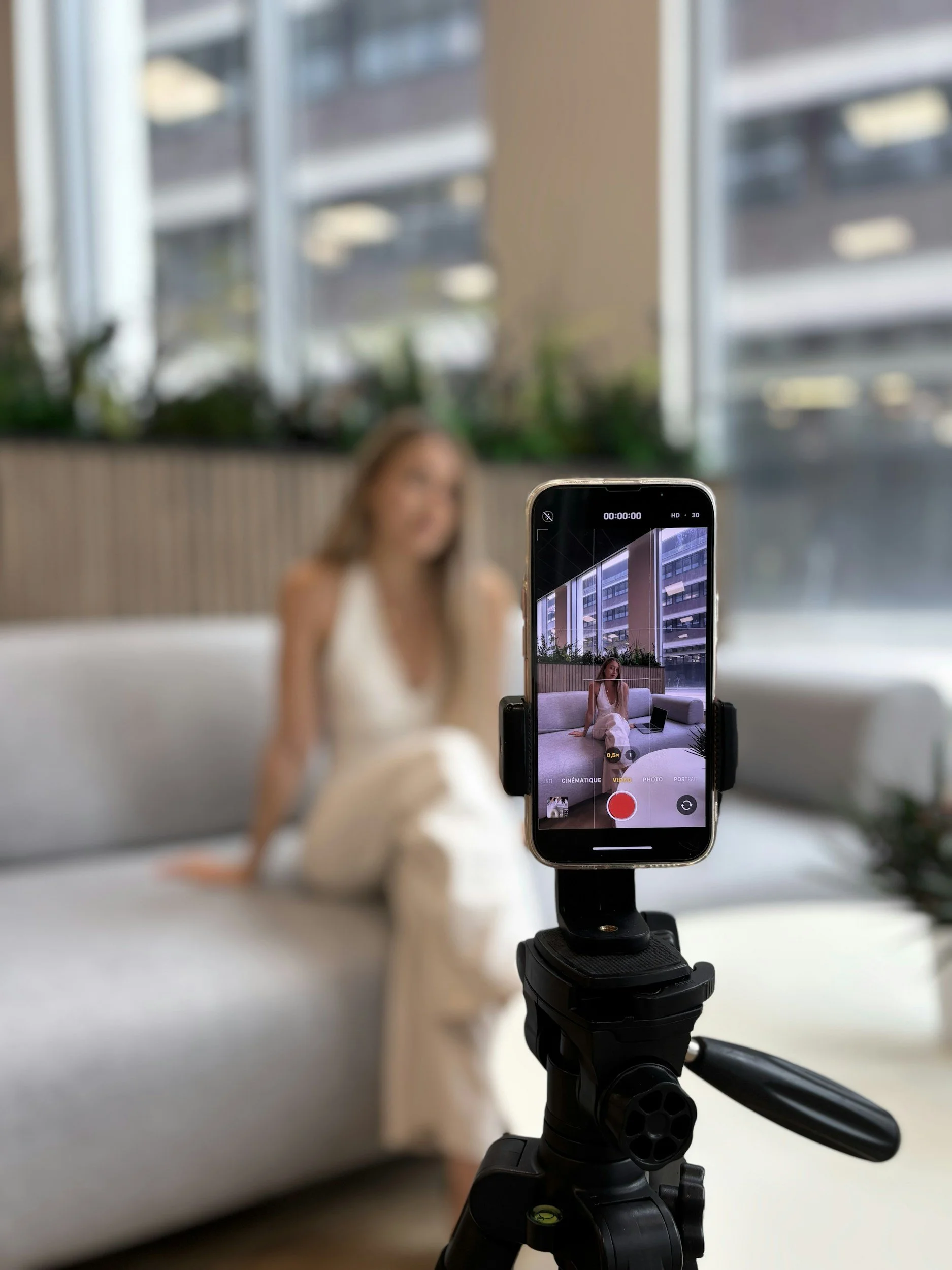 A smartphone mounted on a tripod capturing a woman sitting on a sofa with a laptop, with large windows behind her showing an urban scene.