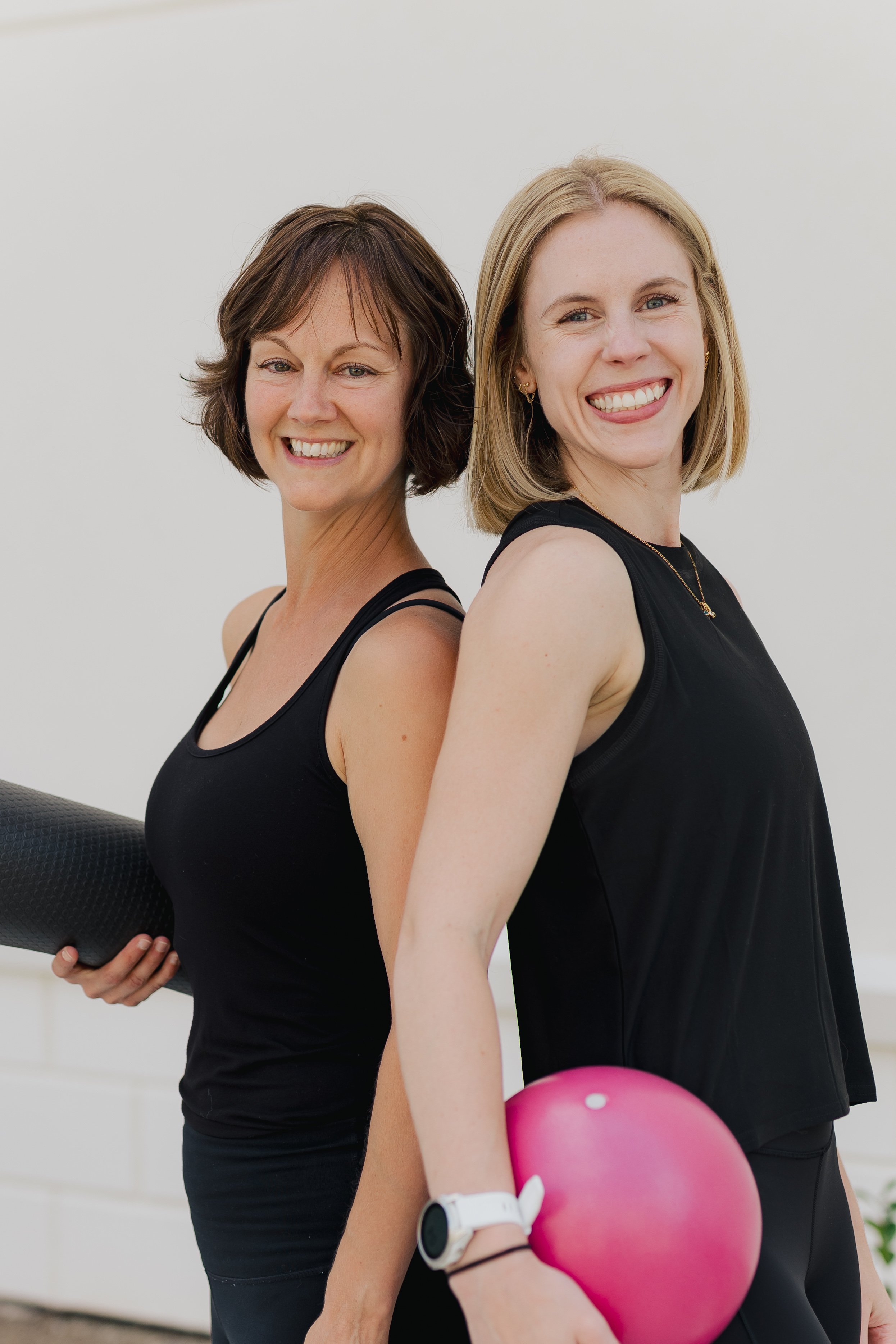Two women in black athletic wear posing back-to-back, with the woman on the right holding a pink exercise ball, in a fitness or gym setting.