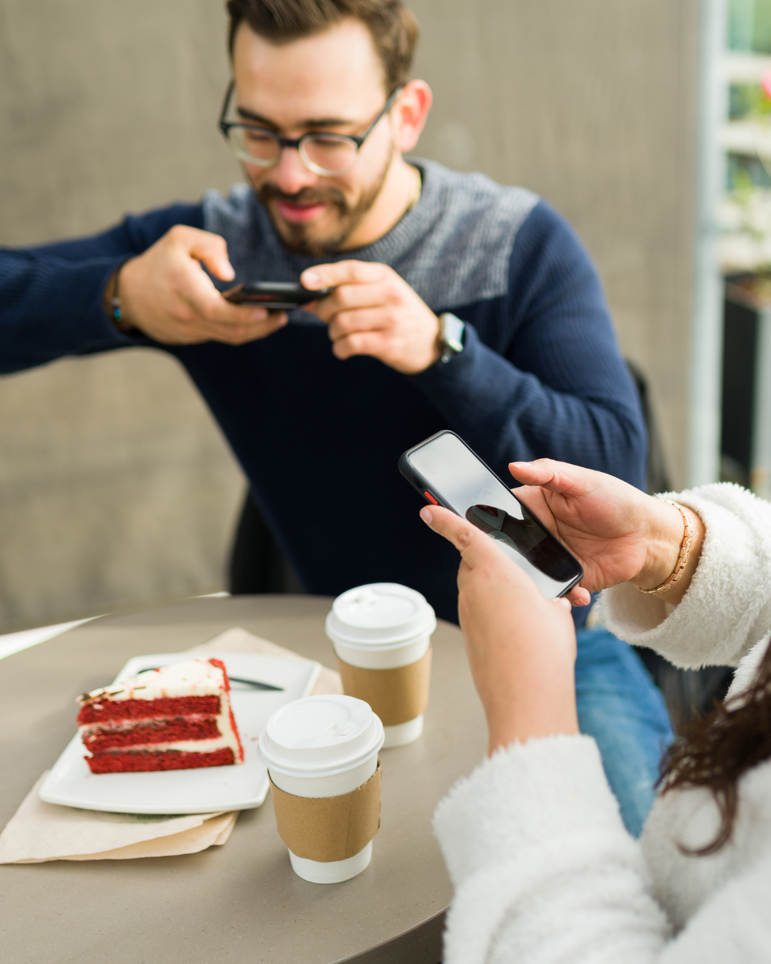 Two people sitting at a table outdoors, both looking at their smartphones. On the table, there are two cups of coffee, a slice of red velvet cake on a white plate, and a napkin.