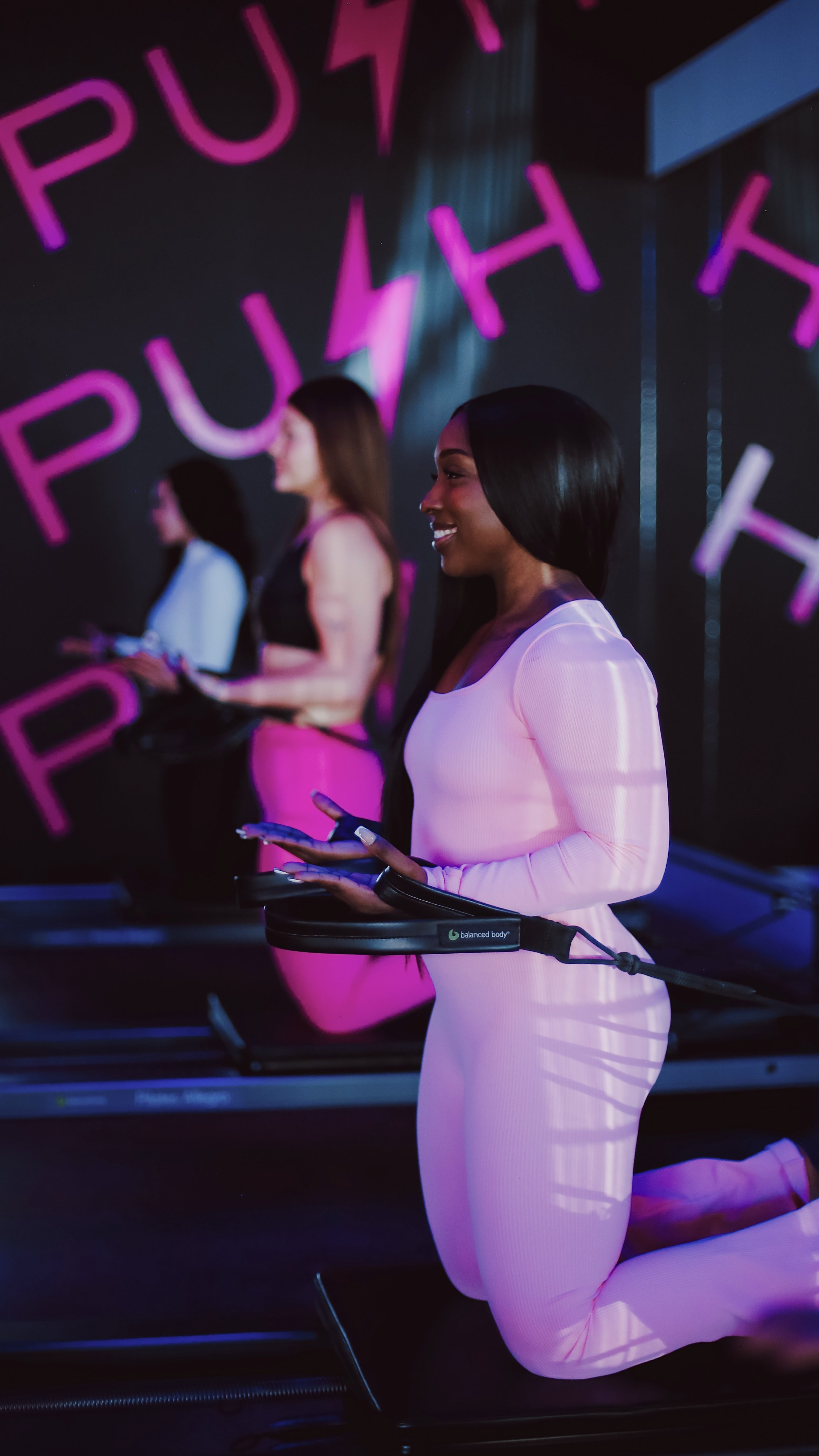 A woman kneeling in a fitness class, wearing a pink workout outfit, with a connected device around her waist, holding a phone in her hands, with a neon-lit background showing the words 'PUSH THE'.