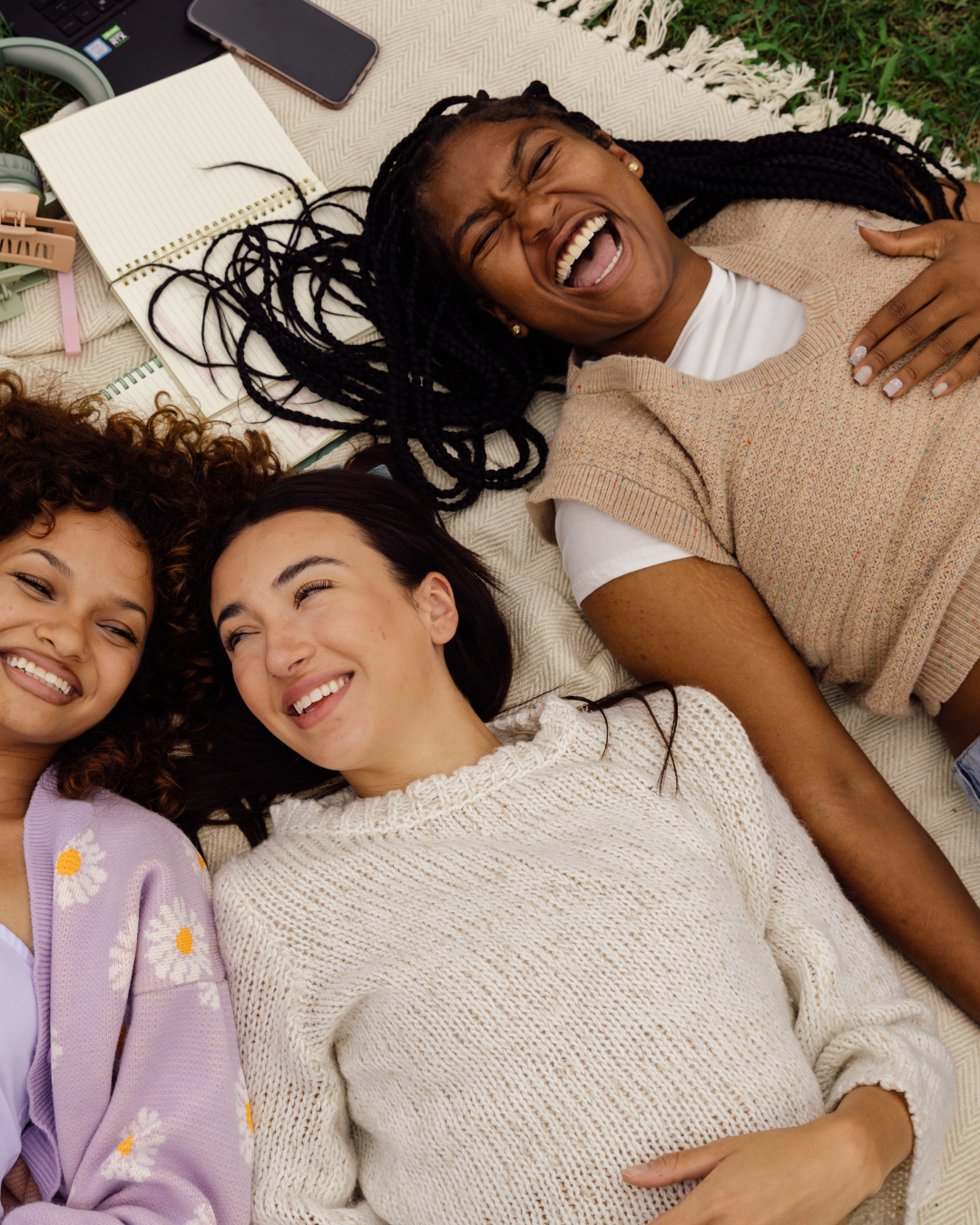 Three women lying on a blanket outdoors, smiling and laughing together, surrounded by notebooks, a phone, and accessories.
