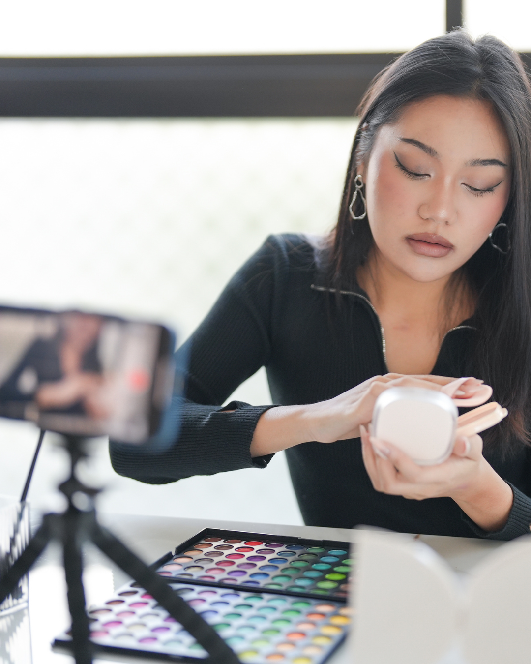 Woman using makeup and filming herself with a smartphone on a tripod, with a colorful eyeshadow palette on the table.