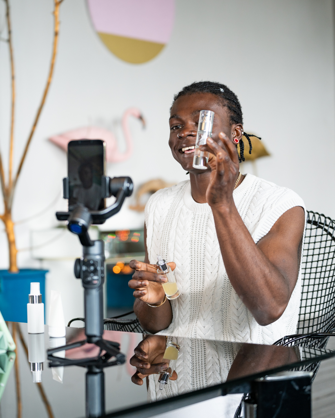 A person recording a skincare product review with a smartphone mounted on a tripod, holding a bottle and a dropper with skincare serum, sitting at a reflective glass table with various skincare bottles.