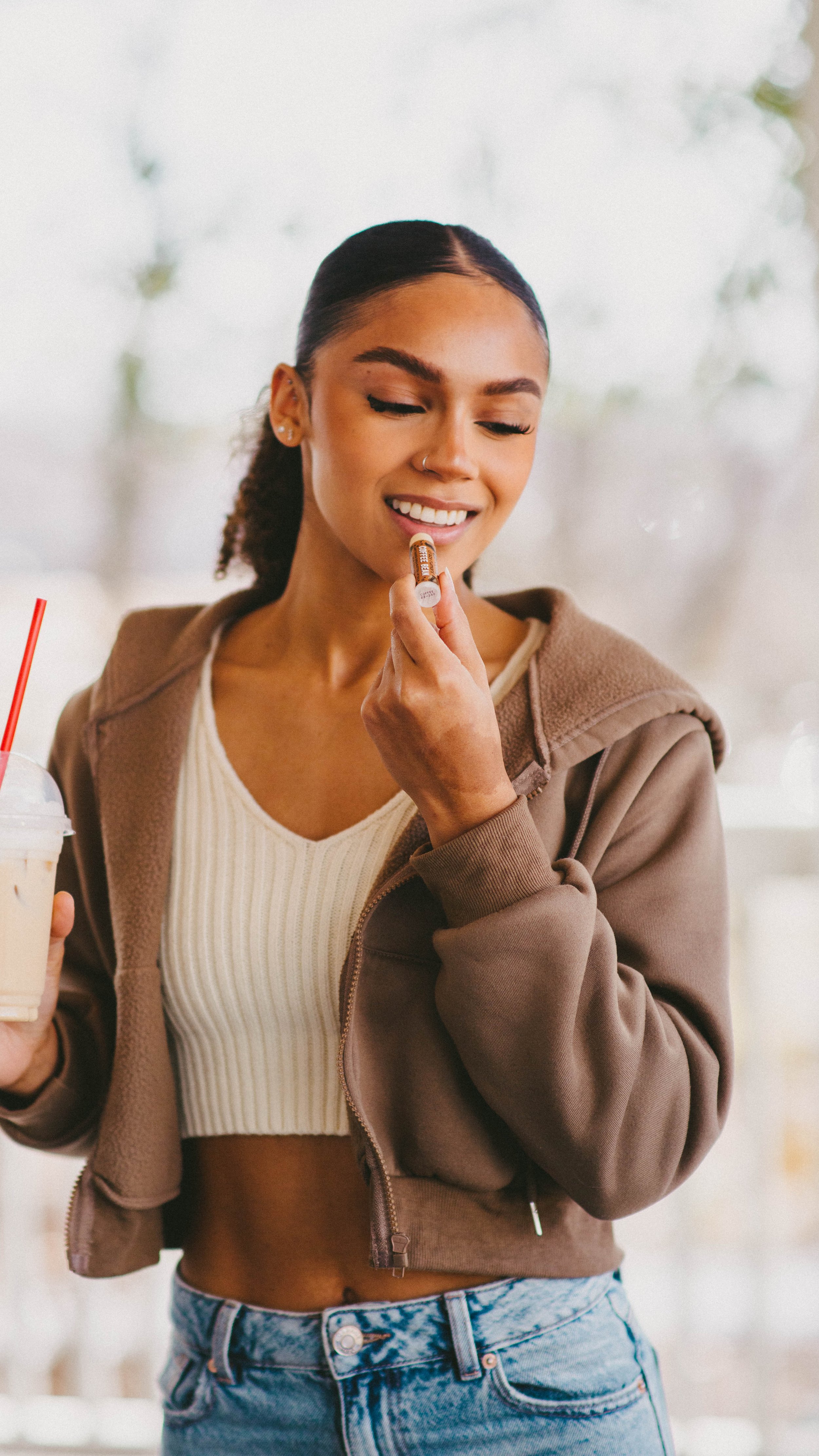 A young woman with dark hair in a ponytail holding a milkshake and applying lipstick in a casual indoor setting.