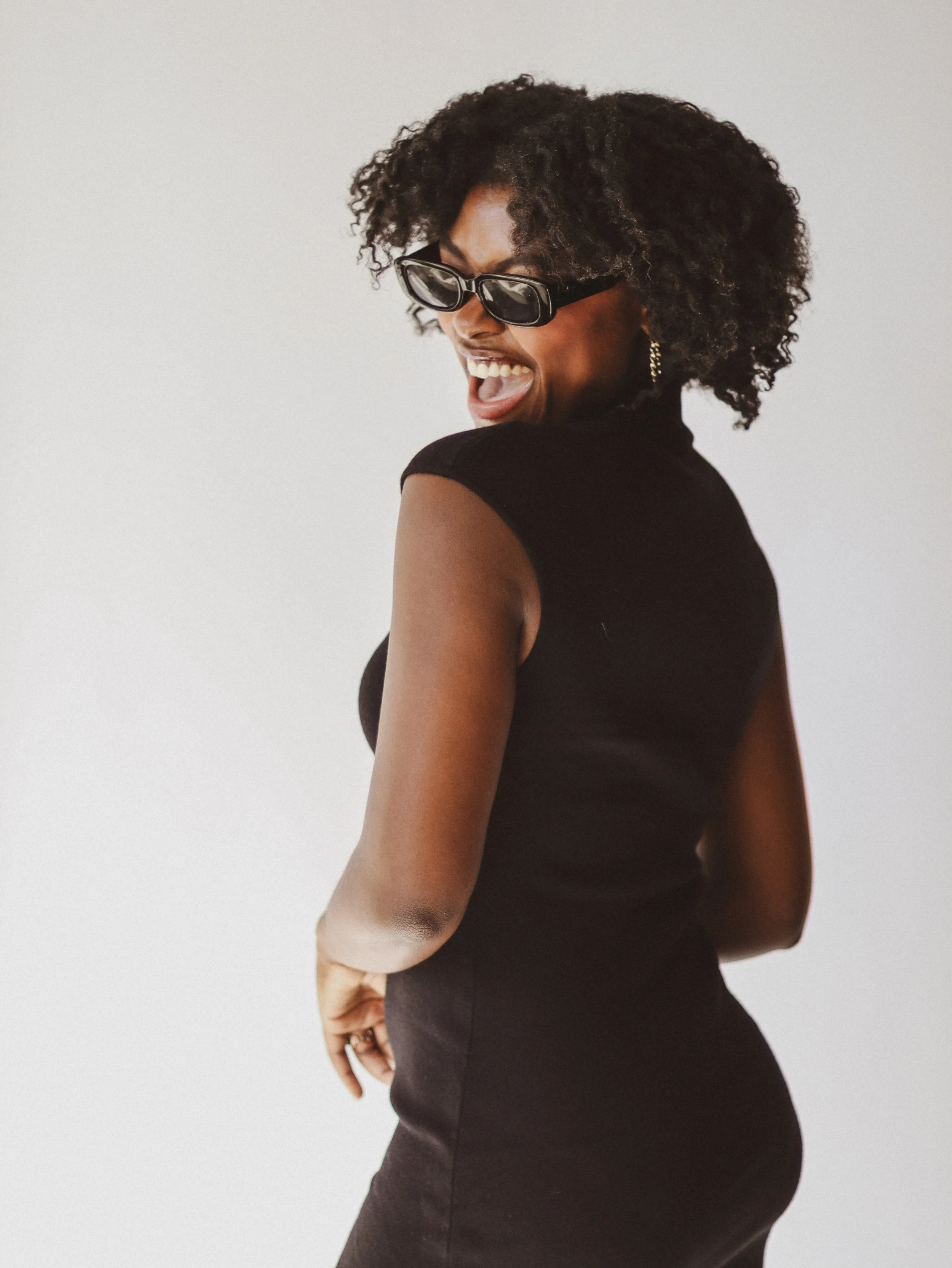 A woman wearing sunglasses, black top, and black skirt, laughing with her mouth open, against a plain white background.