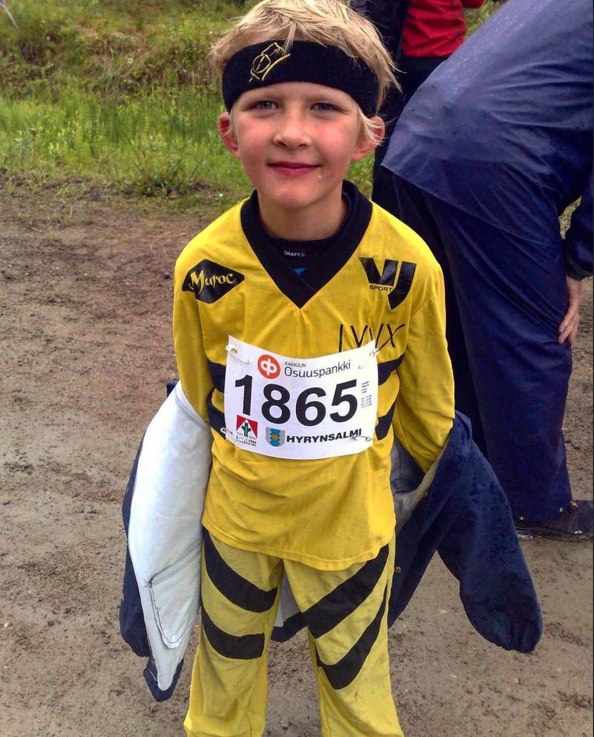 Young boy in yellow sports uniform with race number 1865, holding a jacket and standing outdoors on a dirt path with greenery in the background.