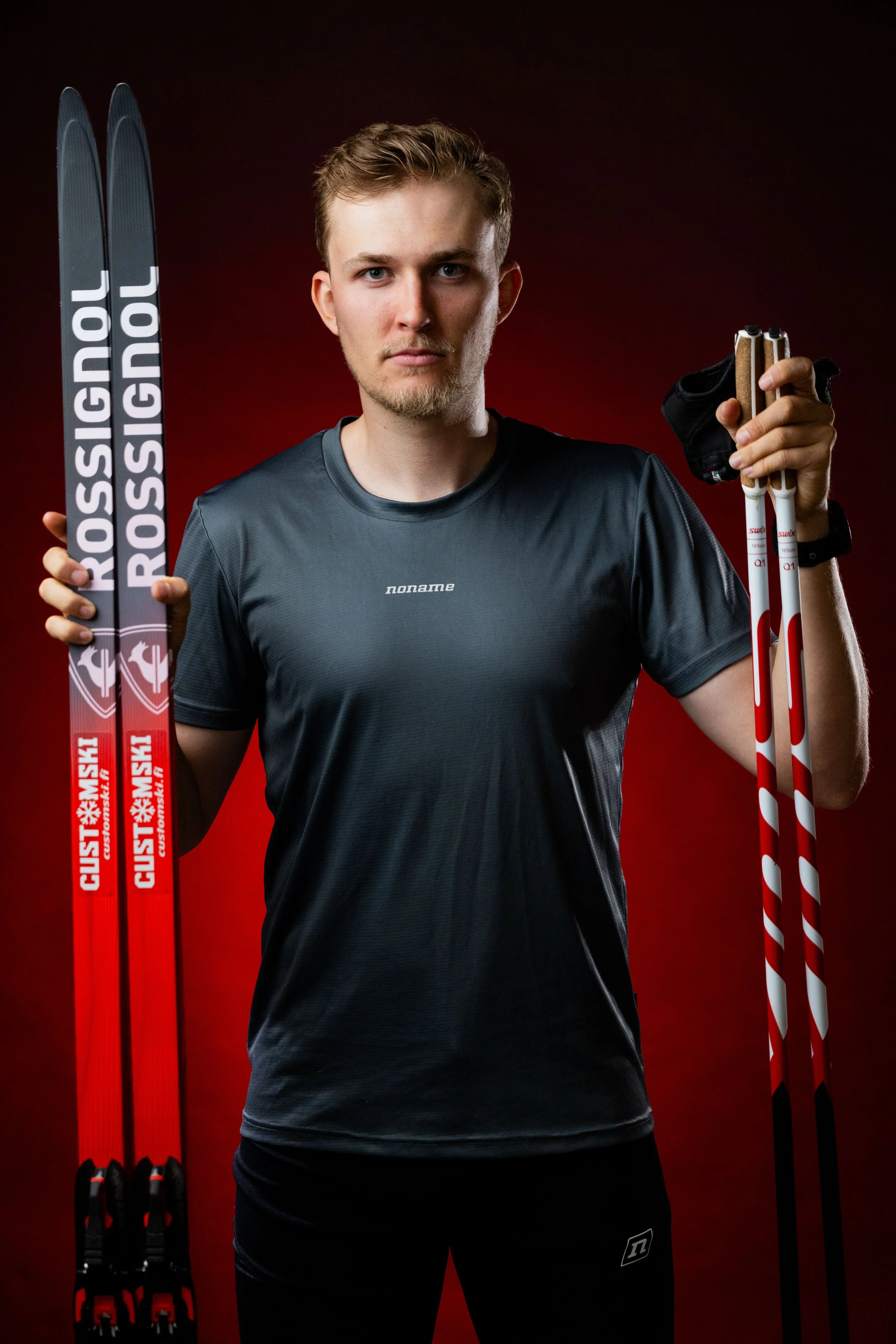 A young male athlete with short, light brown hair and blue eyes, wearing a dark gray sports shirt and black shorts, is holding a pair of red and white cross-country skis on the left and a pair of white and red ski poles on the right, against a dark red background.