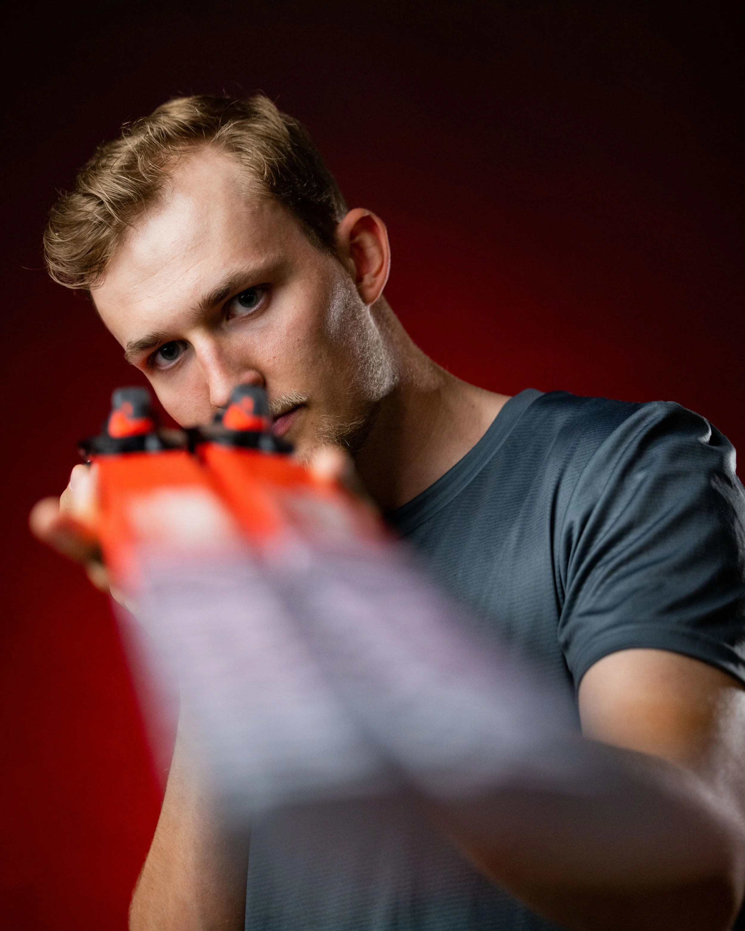 A young man with short, wavy blond hair and blue eyes looking directly into the camera. He is wearing a dark gray shirt and is positioned against a dark red background.
