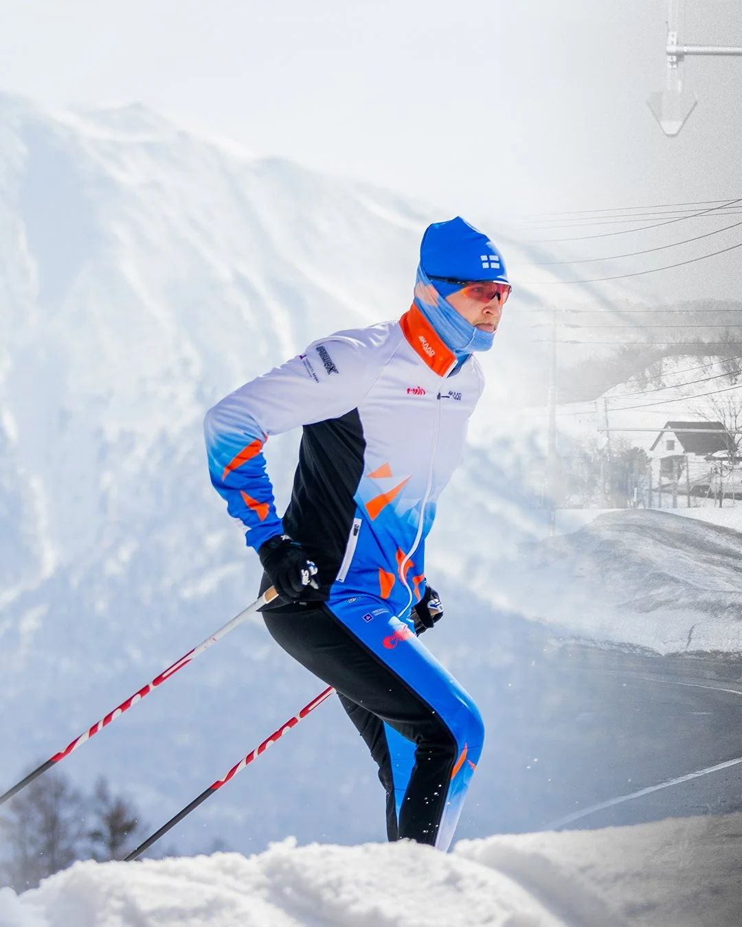 Male cross-country skier wearing a blue, white, black, and orange jacket and matching pants, with a blue hat and red sunglasses, skiing in snowy landscape with mountains and houses in the background.