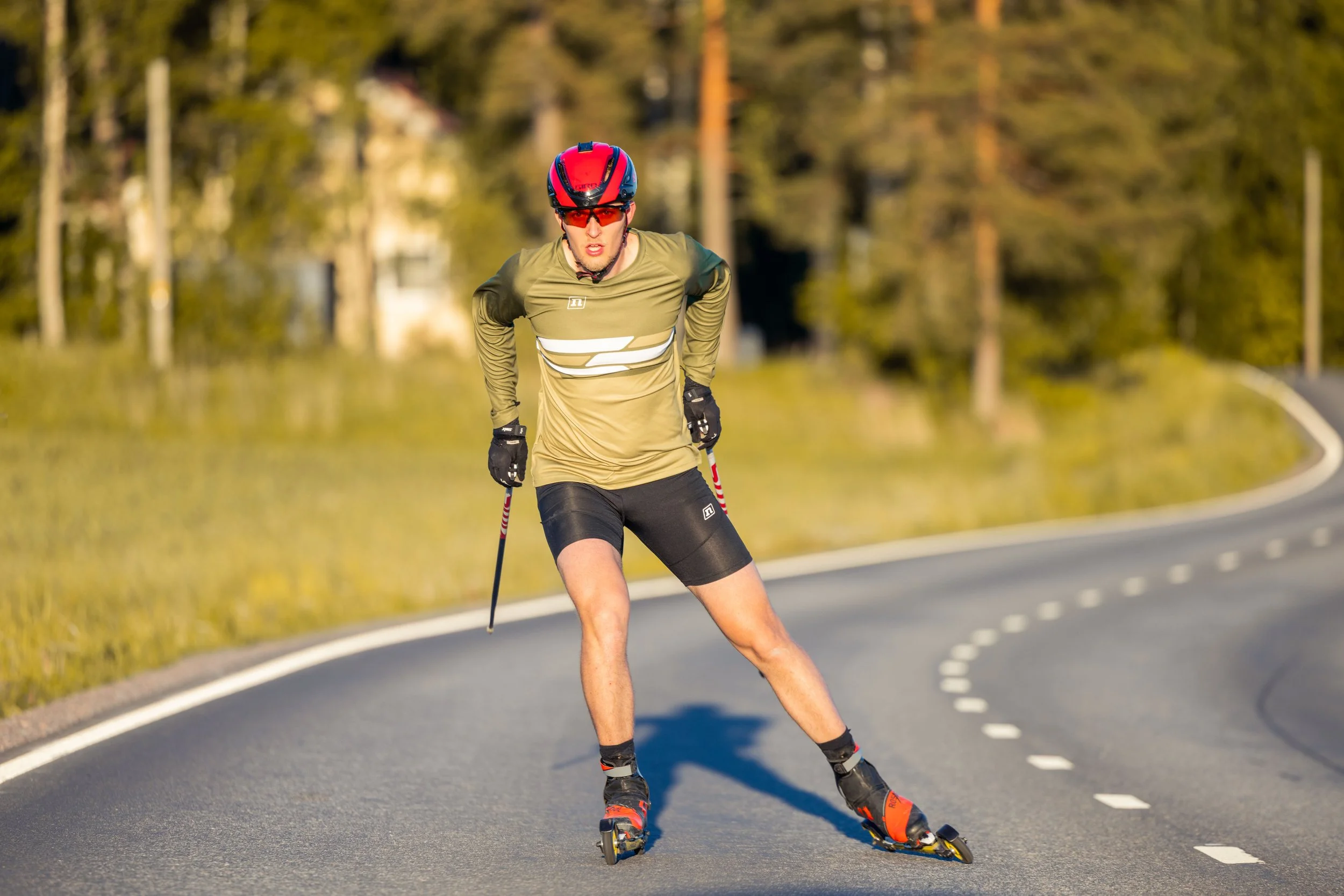 A man wearing a red helmet, sunglasses, and sports clothing is rollerblading on a paved road with white dashed lines, surrounded by trees and greenery.