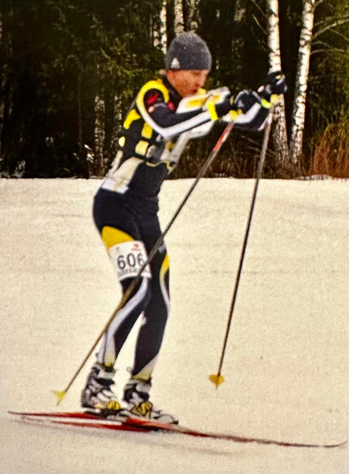 A person in winter sports gear skiing on snow, wearing a black and yellow suit, black gloves, a grey knit hat, and using ski poles, with a forest background.