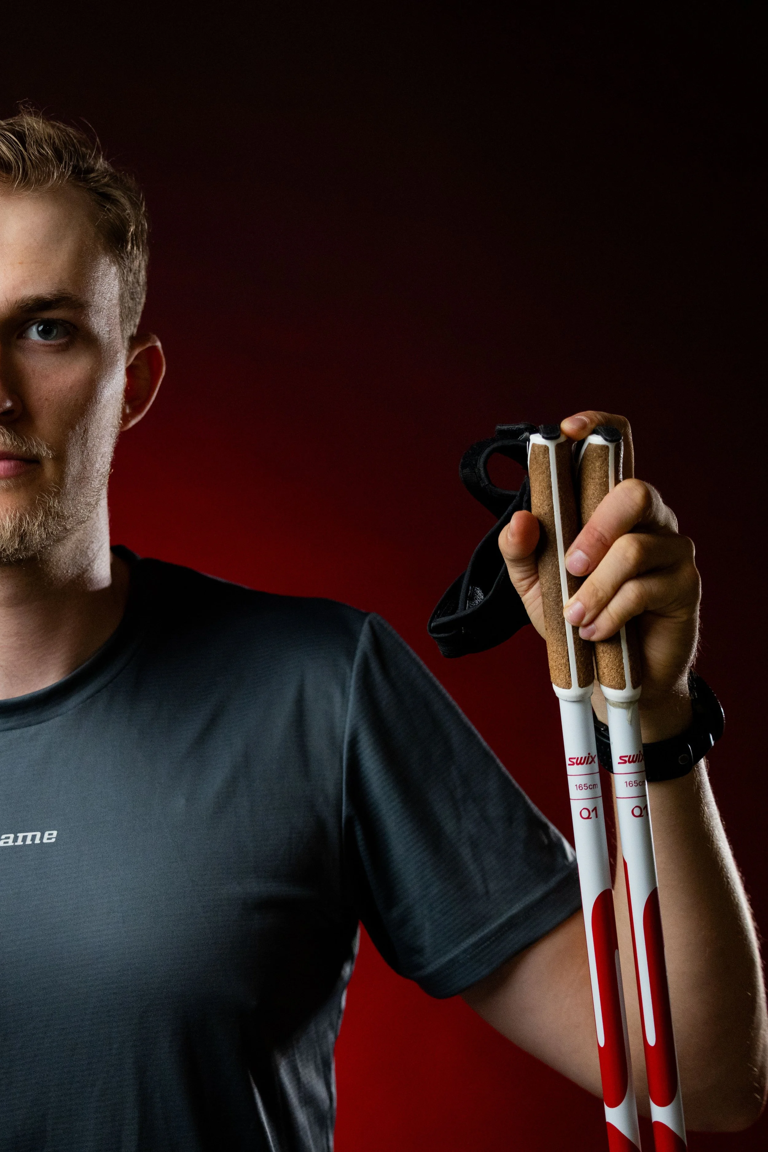 Partial view of a young man holding three white and red cross-country skiing poles, with a dark background.
