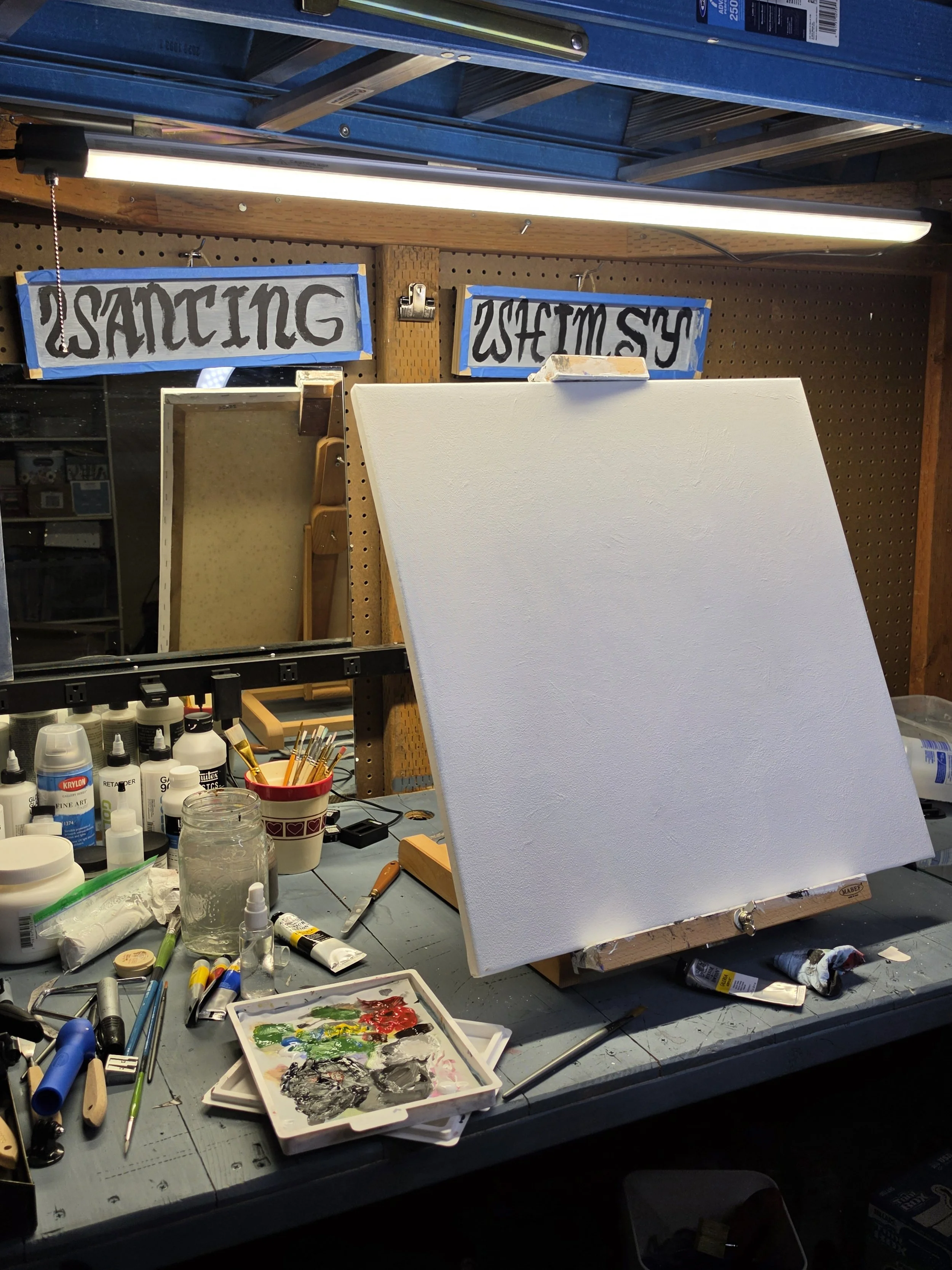 An artist's workspace with a blank canvas, various paints, brushes, and art supplies scattered on the table, illuminated by overhead lighting.
