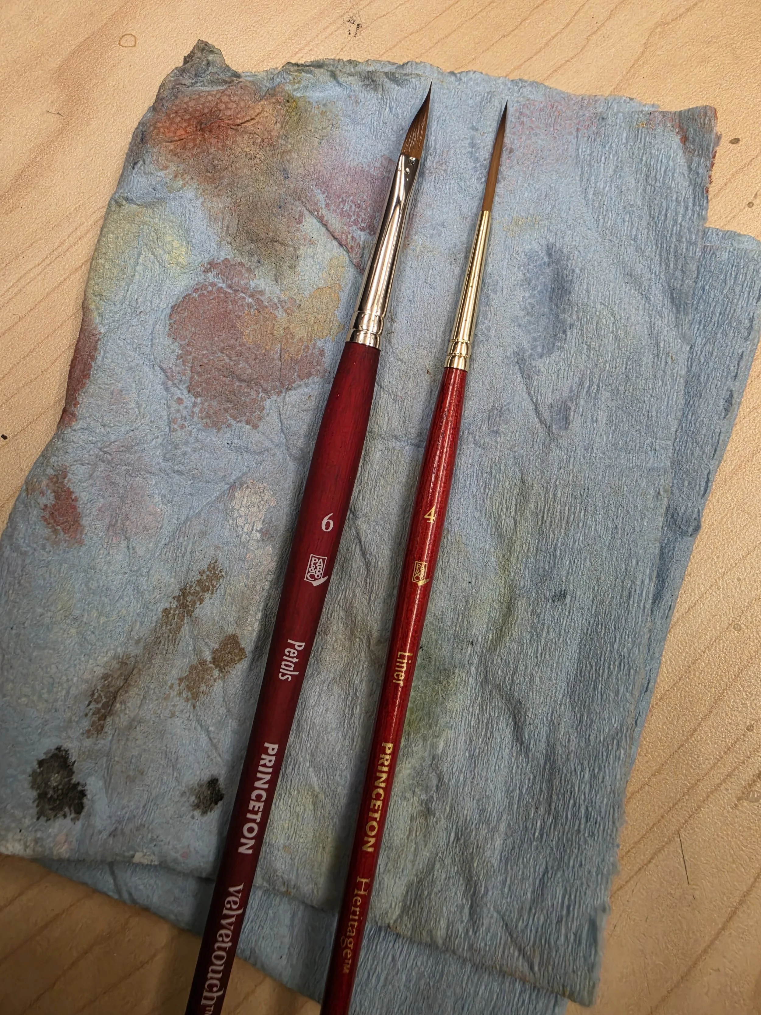 A piece of textured, multicolored paper with two paintbrushes resting on it. The brushes have red handles with gold markings and silver and gold ferrules.